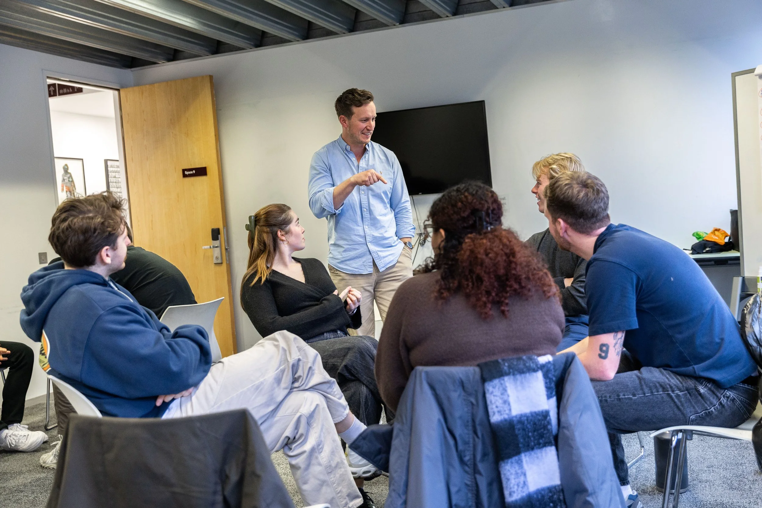 A group of people in a meeting room listening to a man standing and speaking. The man appears to be leading the discussion while others are seated and paying attention.