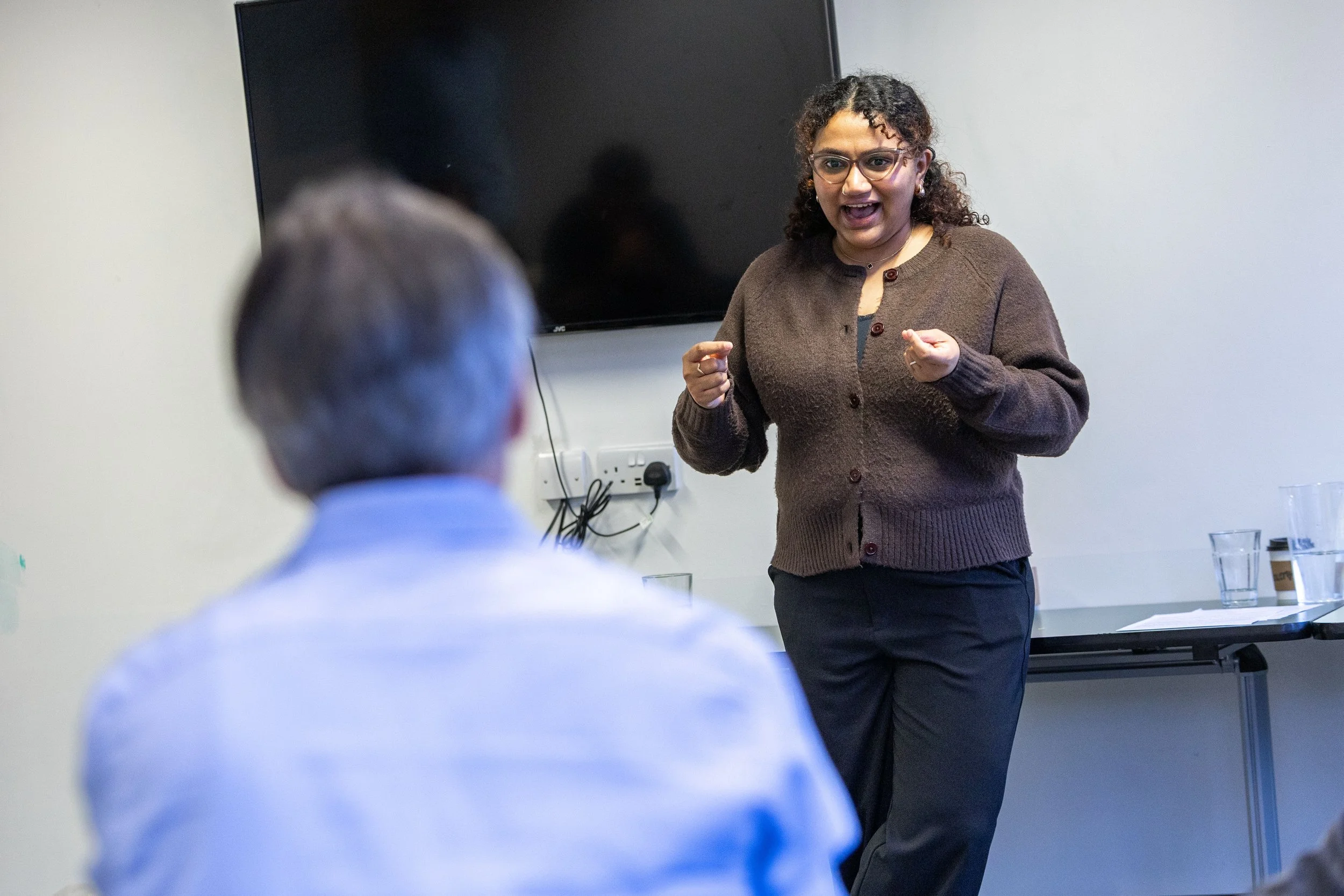A woman speaking and gesturing assertively in front of a seated audience in a conference room.