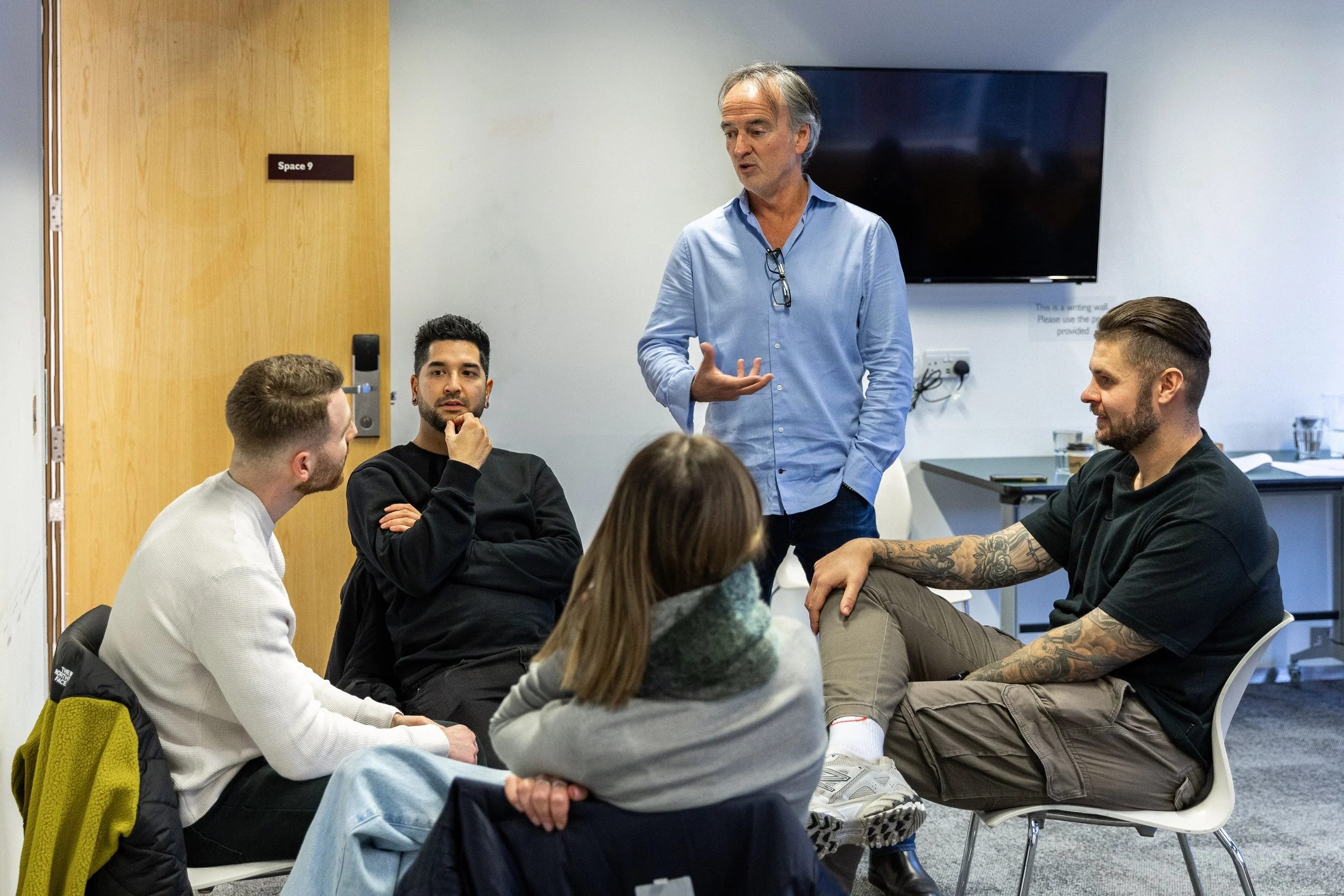 A man stands and speaks to a group of four people seated in a circle in an office meeting room. The man is wearing a light blue shirt, and the seated individuals are listening attentively.