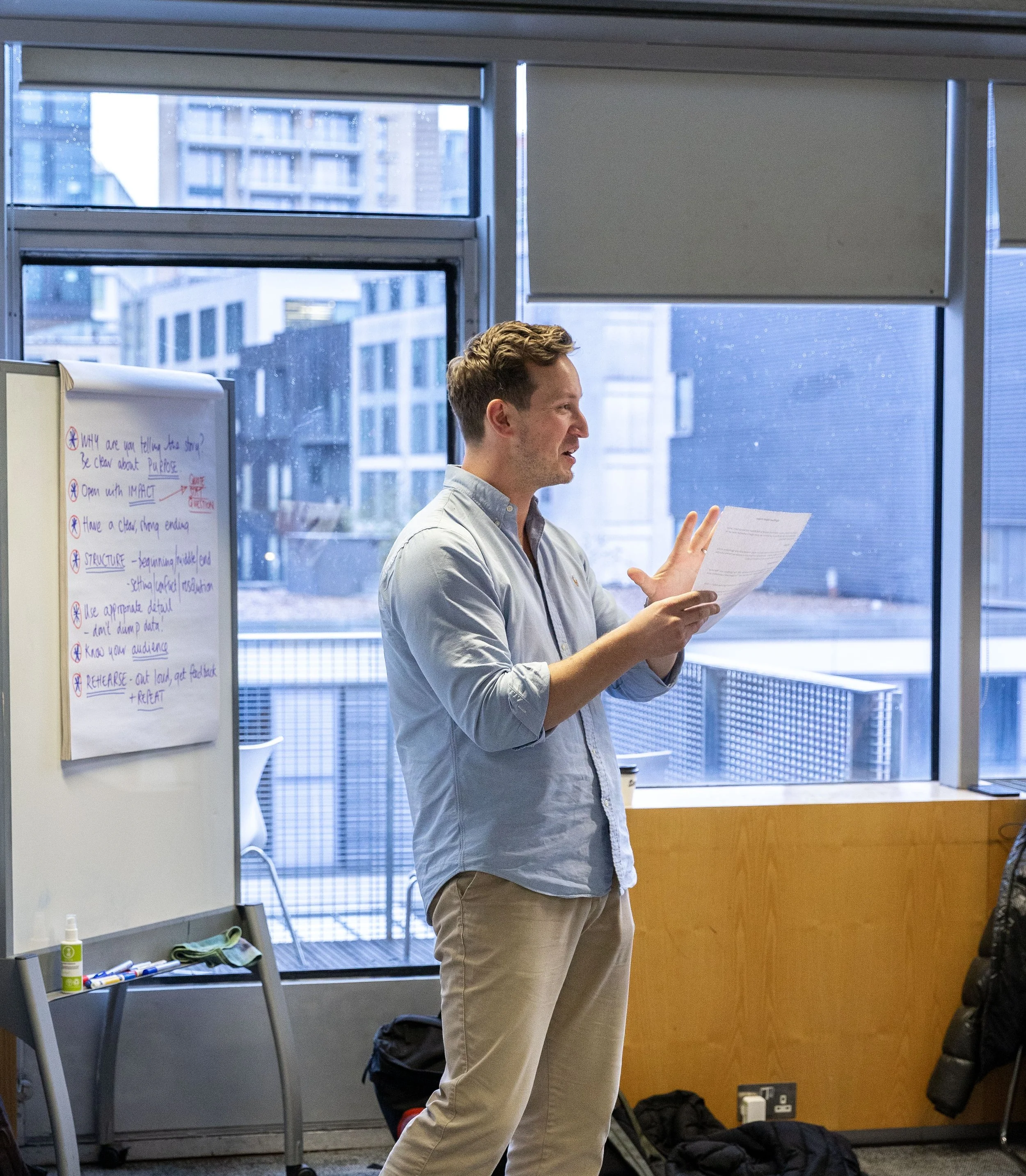 A man in a light blue shirt and beige pants is standing by a window, holding and reading from a paper in an office setting. A whiteboard with writing is visible on the left side of the image.