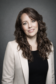A woman with long, wavy brown hair and a light complexion, wearing a white blazer over a black top, smiling against a gray background.