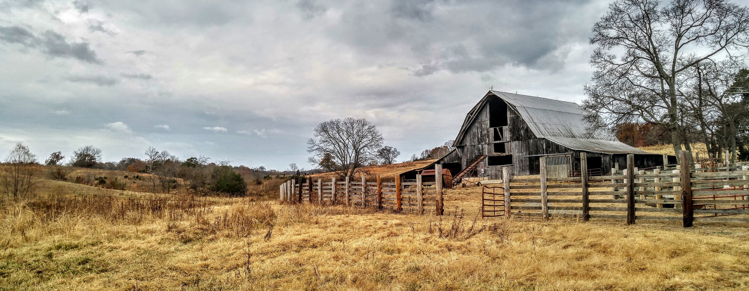 A rustic wooden barn on a hillside with a surrounding wooden fence and leafless trees under a cloudy sky.