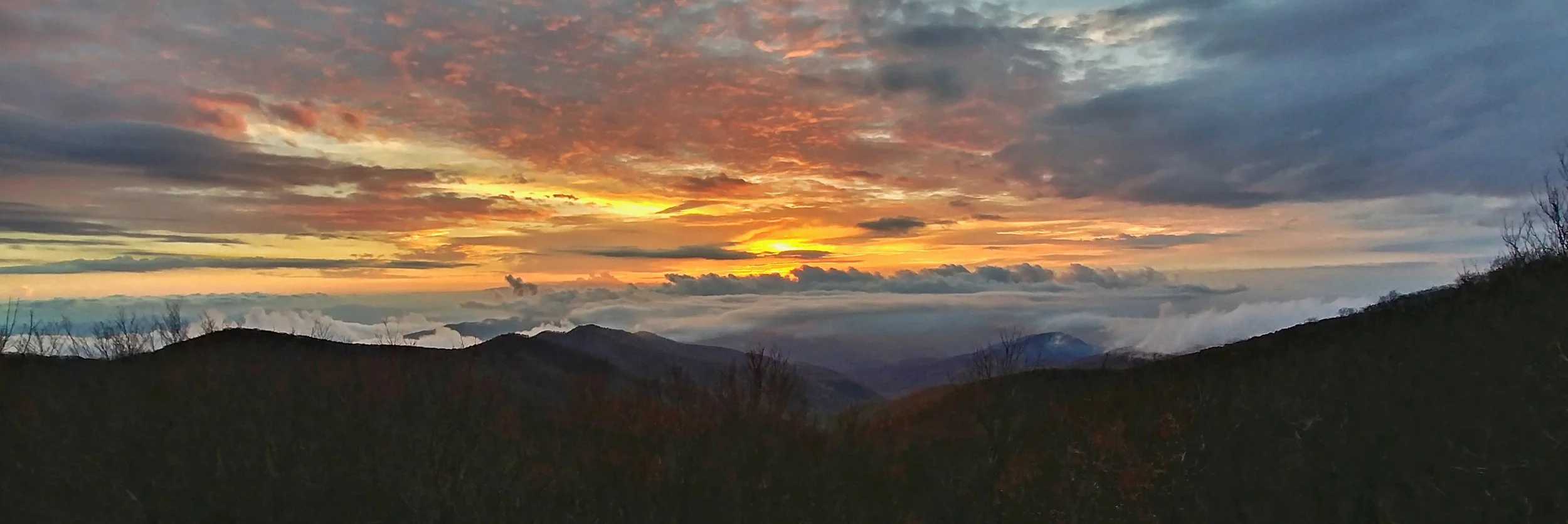 A scenic mountain landscape at sunset with colorful sky and clouds, silhouetted hills, and distant mountains.