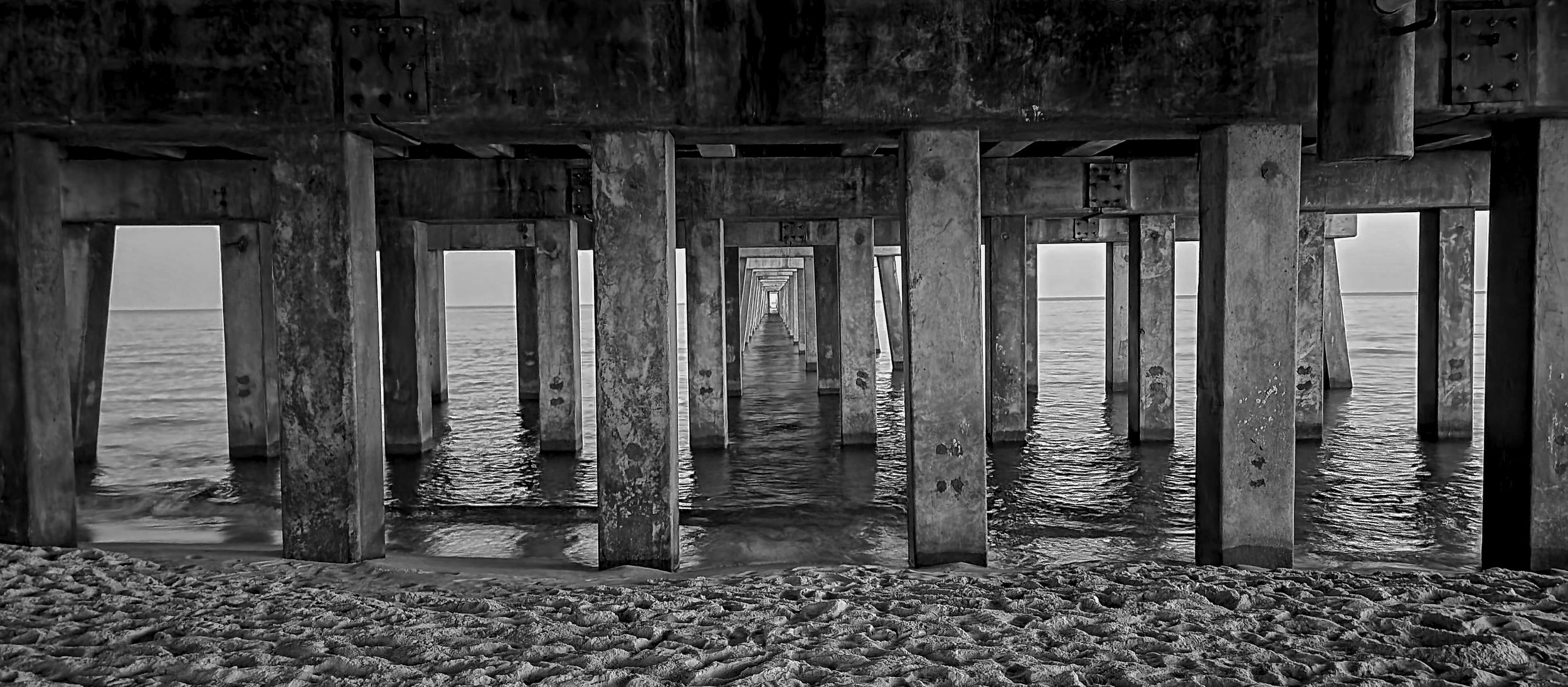 View from underneath a pier at the beach, showing concrete support beams extending into the water, with the ocean and horizon in the background.