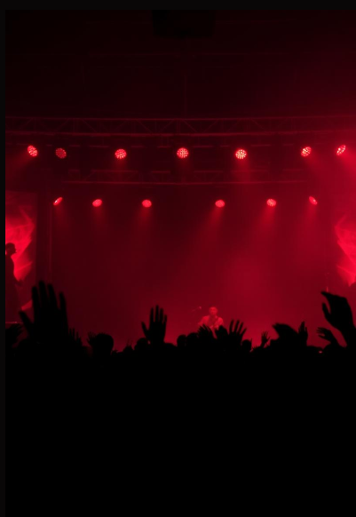 Concert stage with red lighting and silhouettes of audience with raised hands.