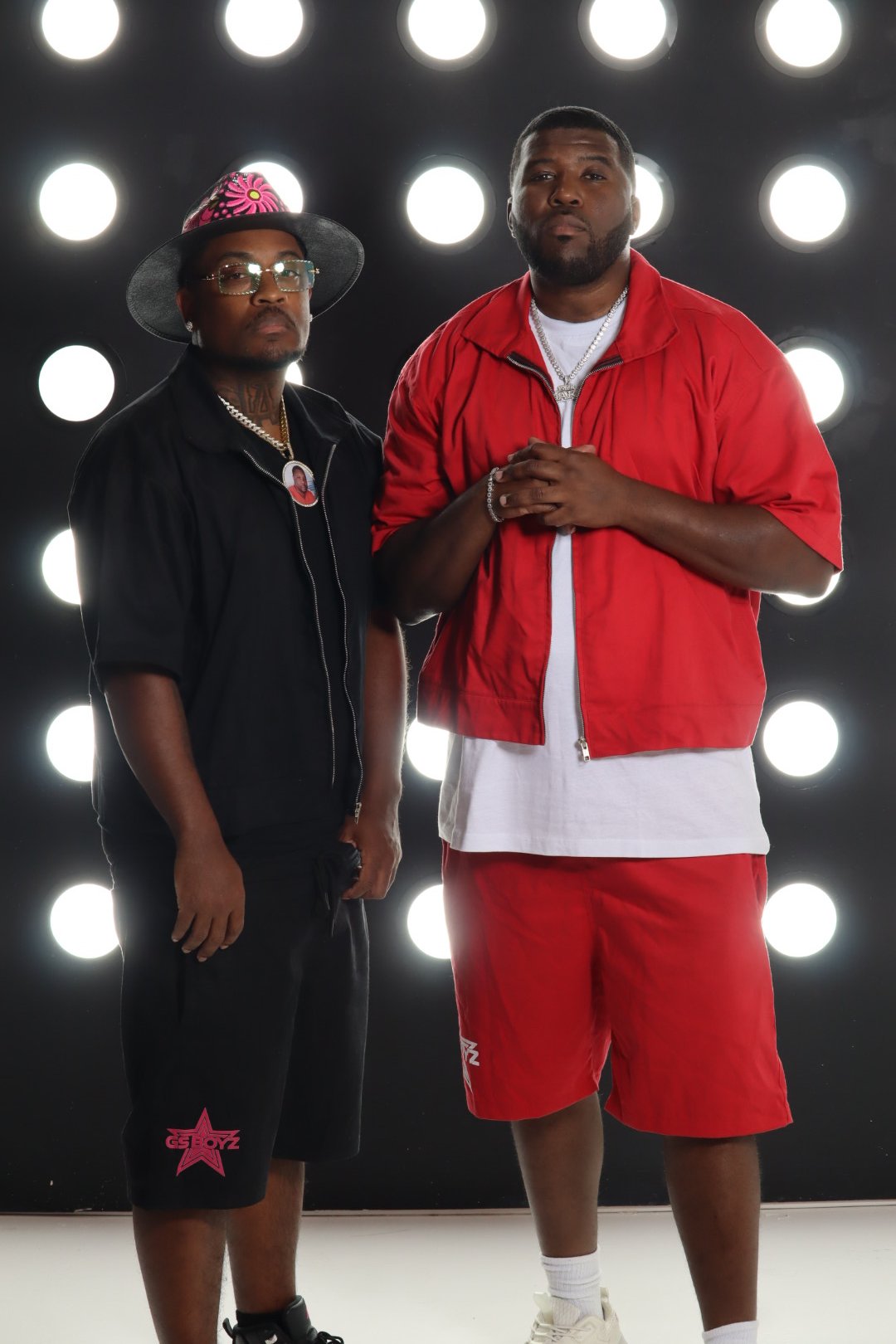 Two men stand in front of a black background illuminated by bright circular lights. One wears a black outfit with a wide-brimmed hat, glasses, and a chain, while the other wears a red jacket over a white shirt and red shorts, each with jewelry and a serious expression.