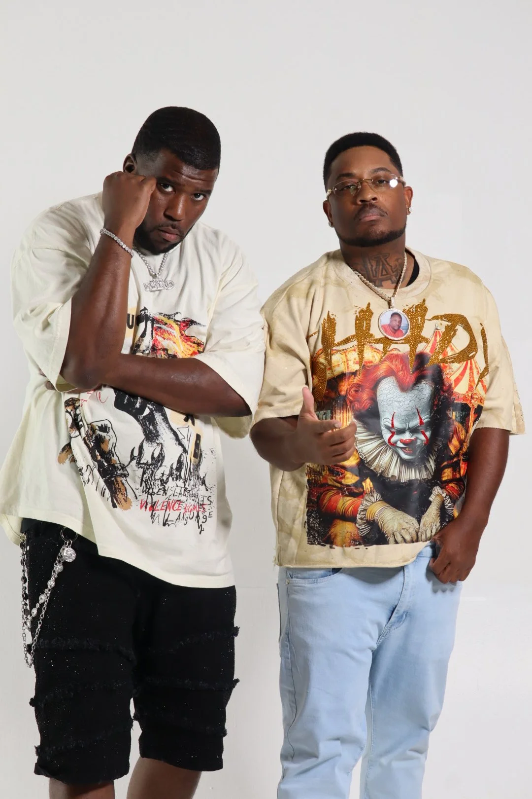 Two men standing against a plain white background, both wearing graphic T-shirts with horror movie characters and making expressive gestures, one with a fist near his head and the other giving a thumbs-up.