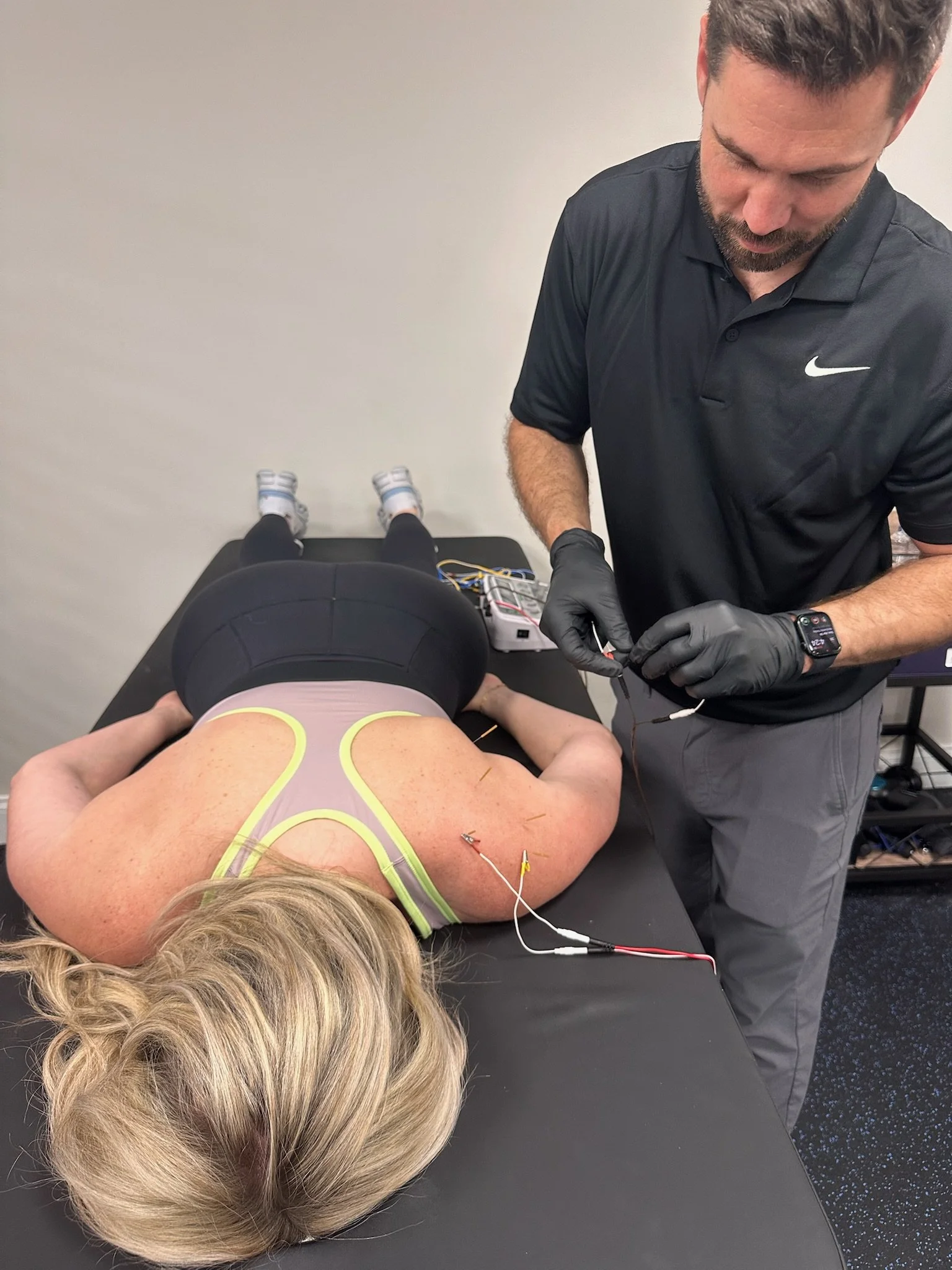 A woman lying face down on a table with electrodes attached to her back, receiving electrotherapy treatment. A man wearing gloves is preparing or adjusting the equipment as part of the therapy session.