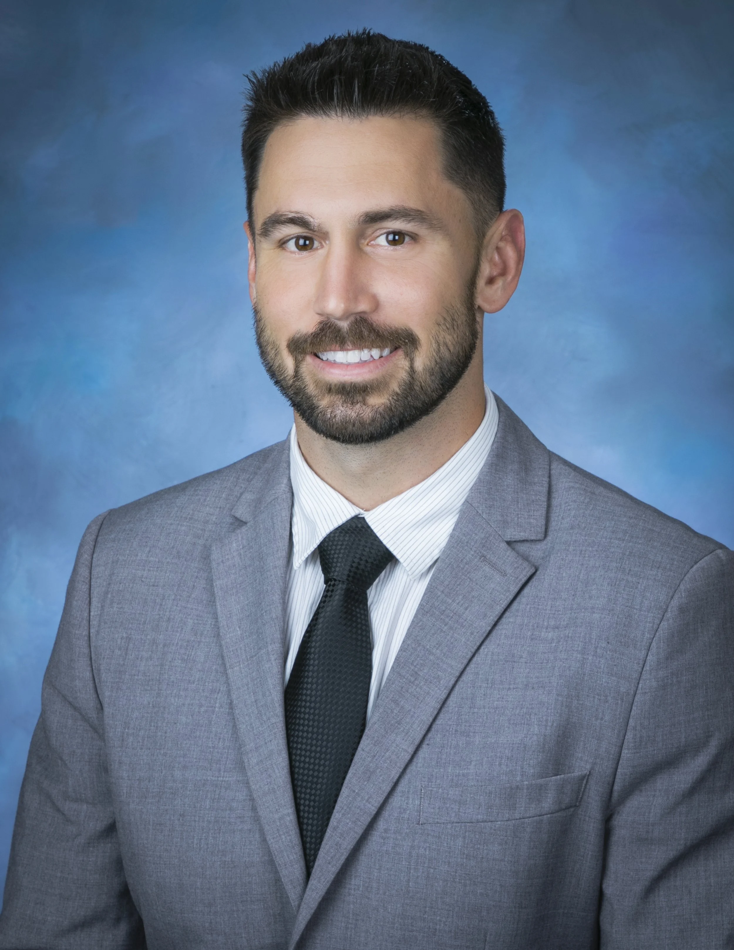 A man in a gray suit with a white shirt and black tie, smiling against a blue background.