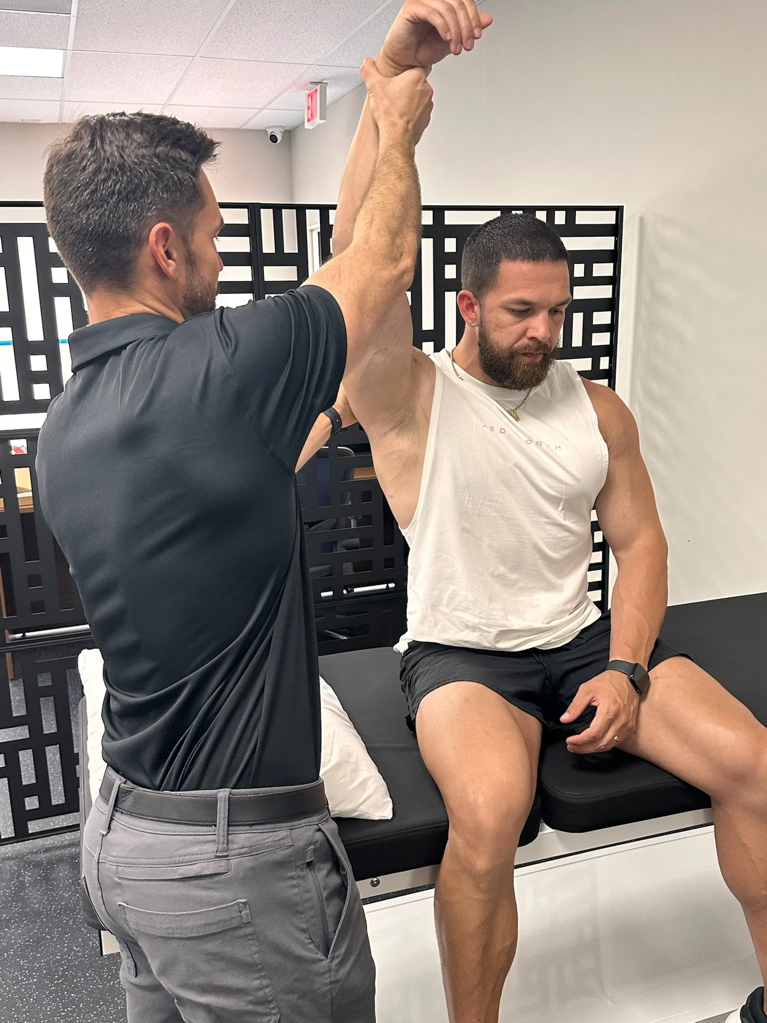 A physical therapist is helping a man with a beard stretch his arm while seated on a treatment table in a clinical setting.