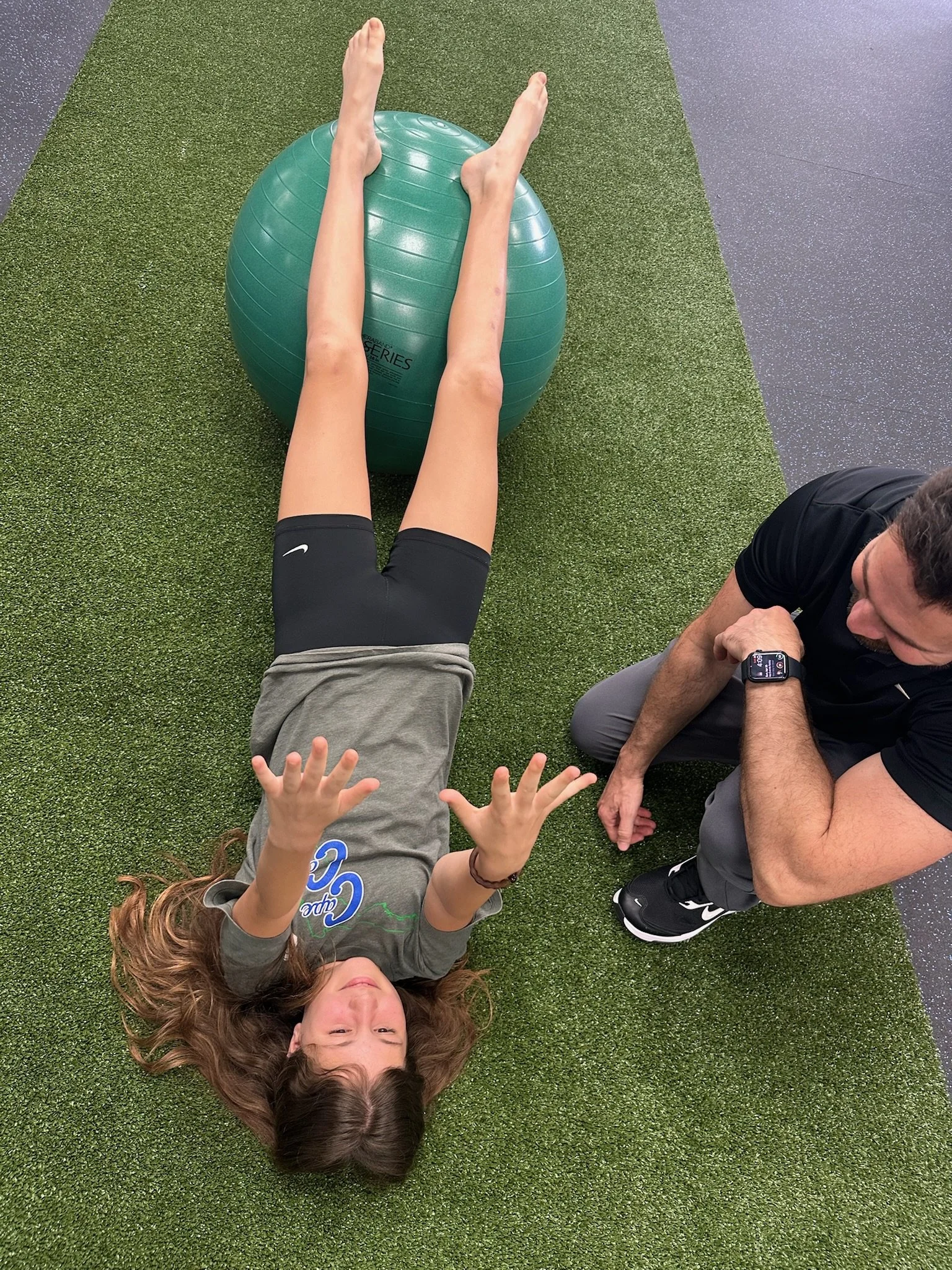 A woman with long, wavy brown hair is lying on her back on artificial green turf. Her eyes are closed, and she is smiling. She is wearing a gray T-shirt with blue and green graphics. Her arms are raised, and her hands are making peace signs. Another person, wearing a black T-shirt, gray pants, and Nike sneakers, is kneeling next to her, looking at her and holding a smartwatch.