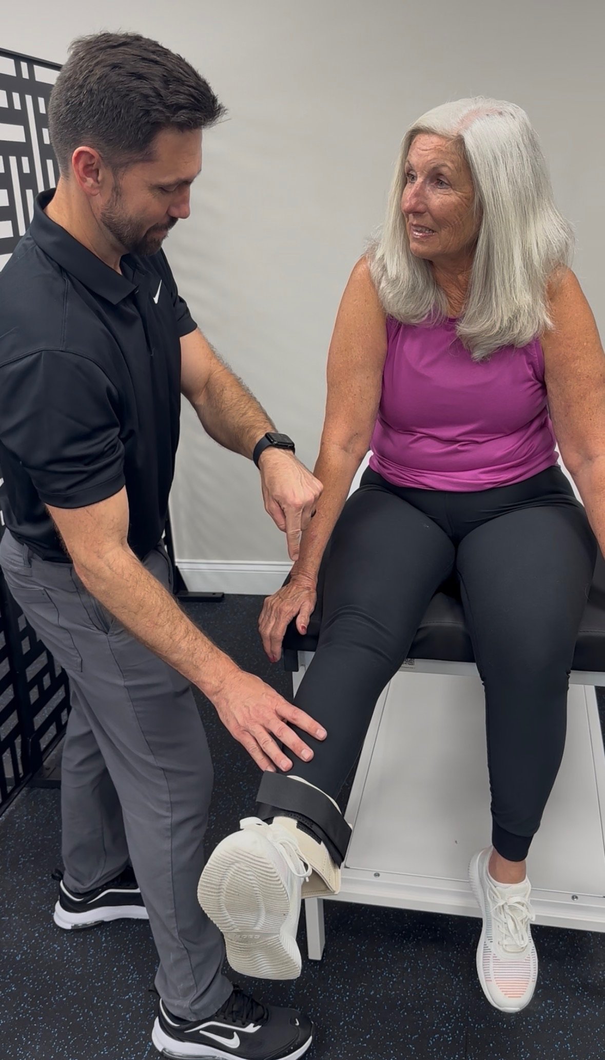A woman sitting on a medical examination table with her leg elevated, while a healthcare professional checks her knee, likely during a physical therapy session.