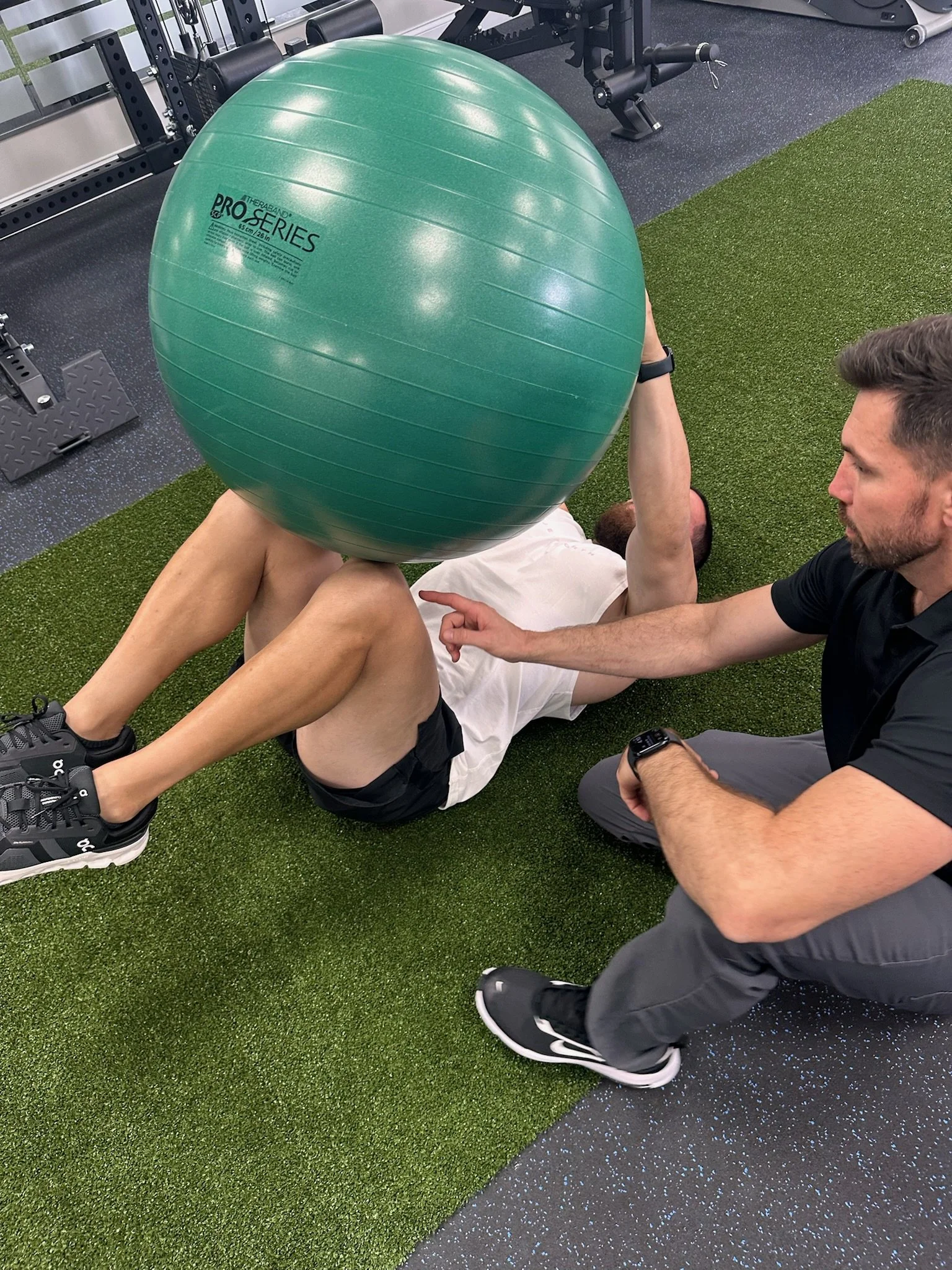 A person lying on the ground holding an exercise ball over their face while another person, a physical therapist , supports and instructs them in a gym with artificial turf and workout equipment.
