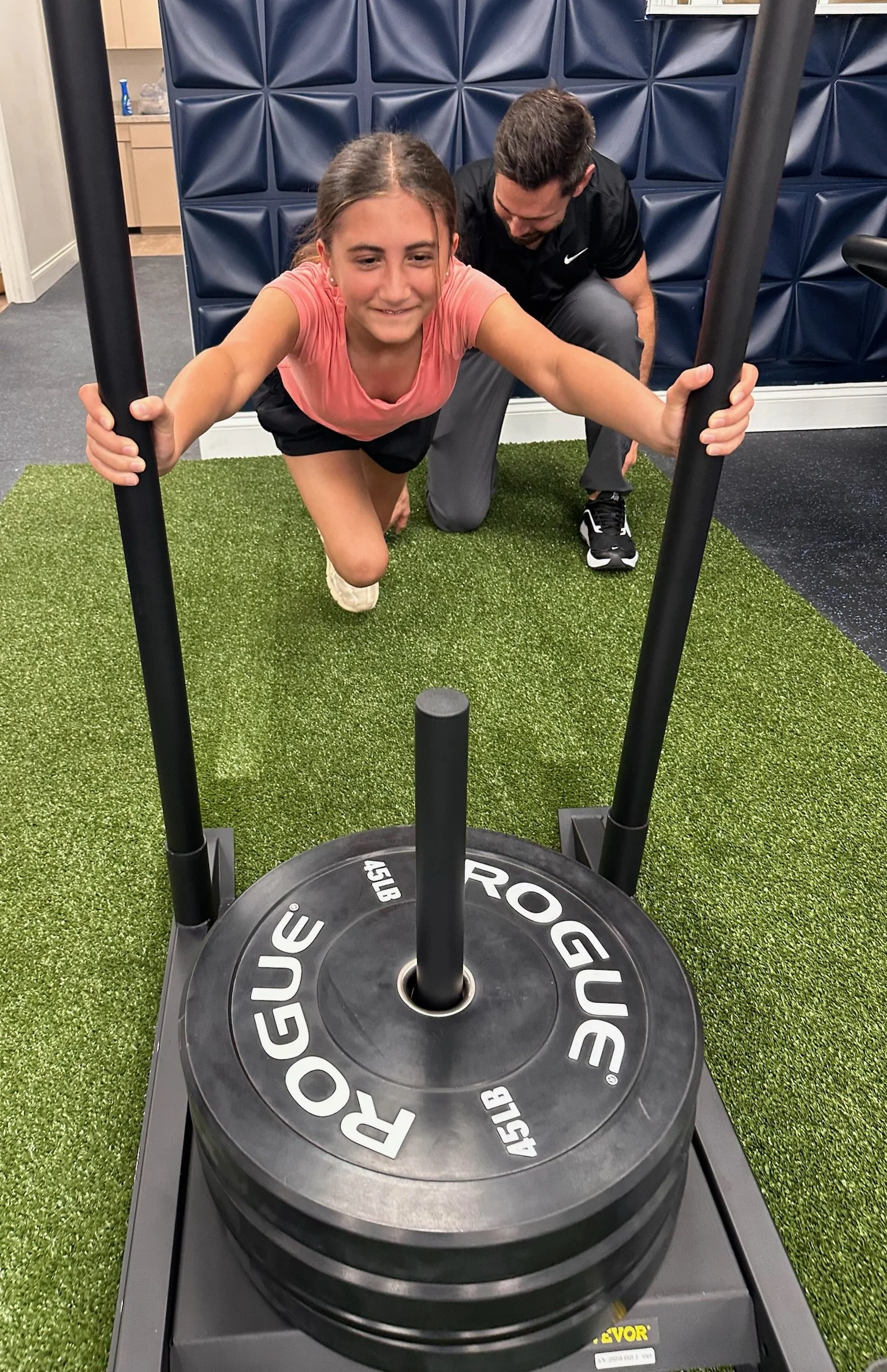 A young girl in a pink shirt and black shorts is doing lunges on a green turf flooring in a gym; a man in a black shirt and gray pants is coaching her from behind.