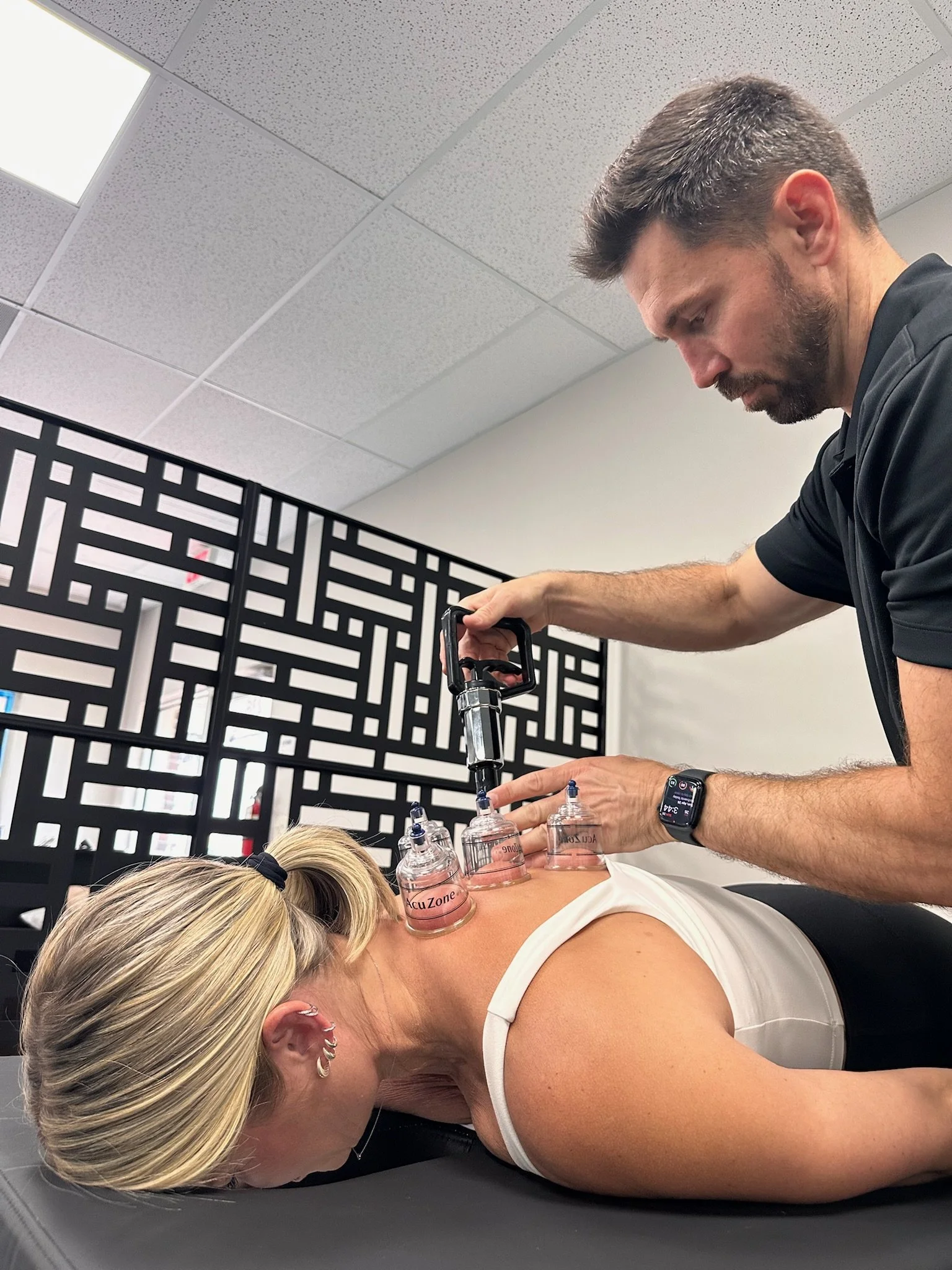A woman lying face down on a treatment table receiving cupping therapy with multiple suction cups on her back, while a practitioner applies a device to enhance the treatment, in a clinical setting.