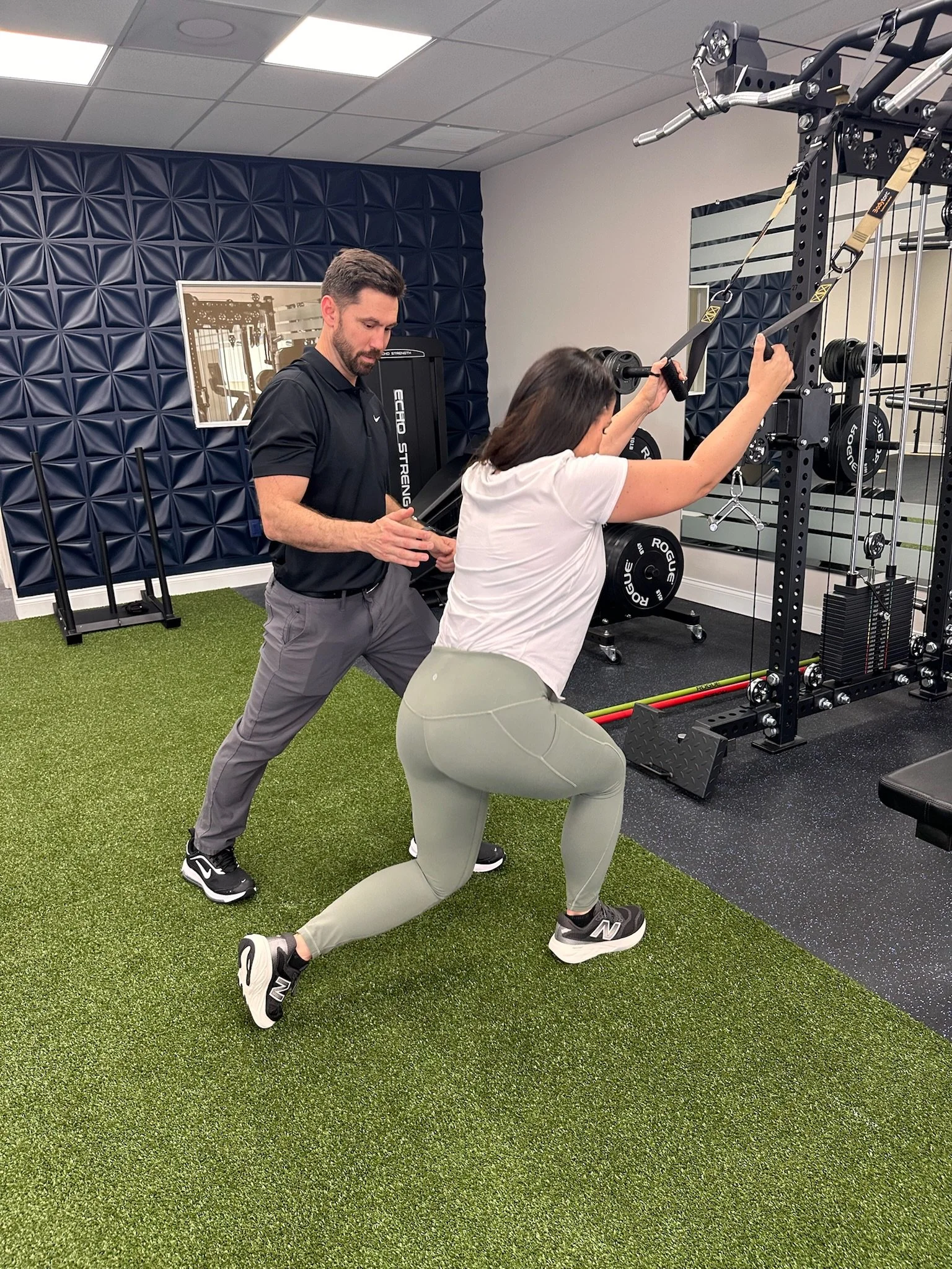 A woman doing a squat exercise with resistance bands while a physical therapist instructs her in a gym with blue padded walls, weight plates, and gym equipment.