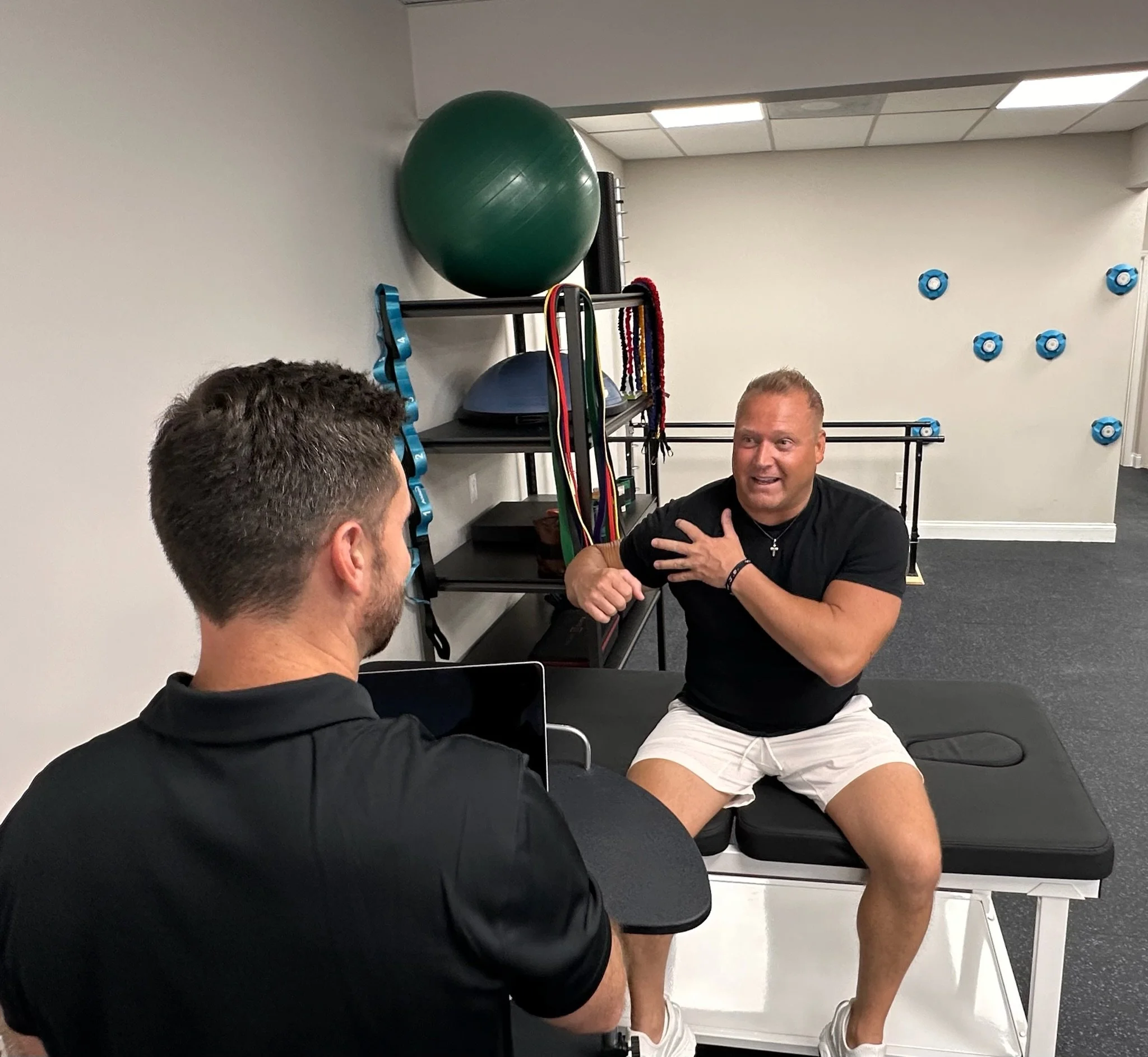 A man sitting on a chair in a gym, with his top half visible, smiling while looking at two men talking to him, with a storage rack in the background holding a green exercise ball, resistance bands, and other equipment, against a wall with blue climbing holds.