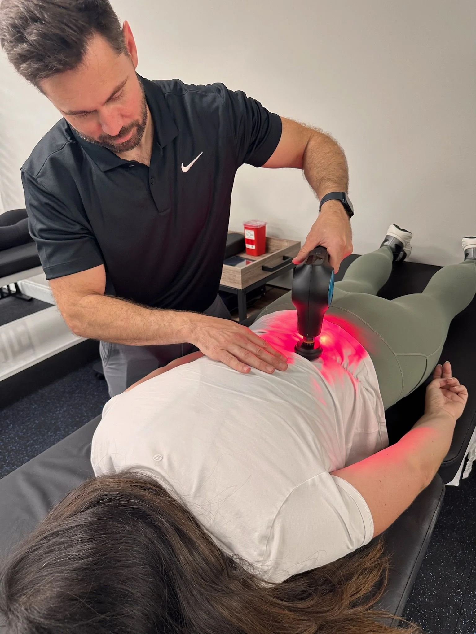 A person lying face down on a massage table receiving a therapy with a device emitting red light. The therapist, wearing a black Nike polo shirt, is holding the device over the person's lower back. The setting appears to be a clinic or therapy room.