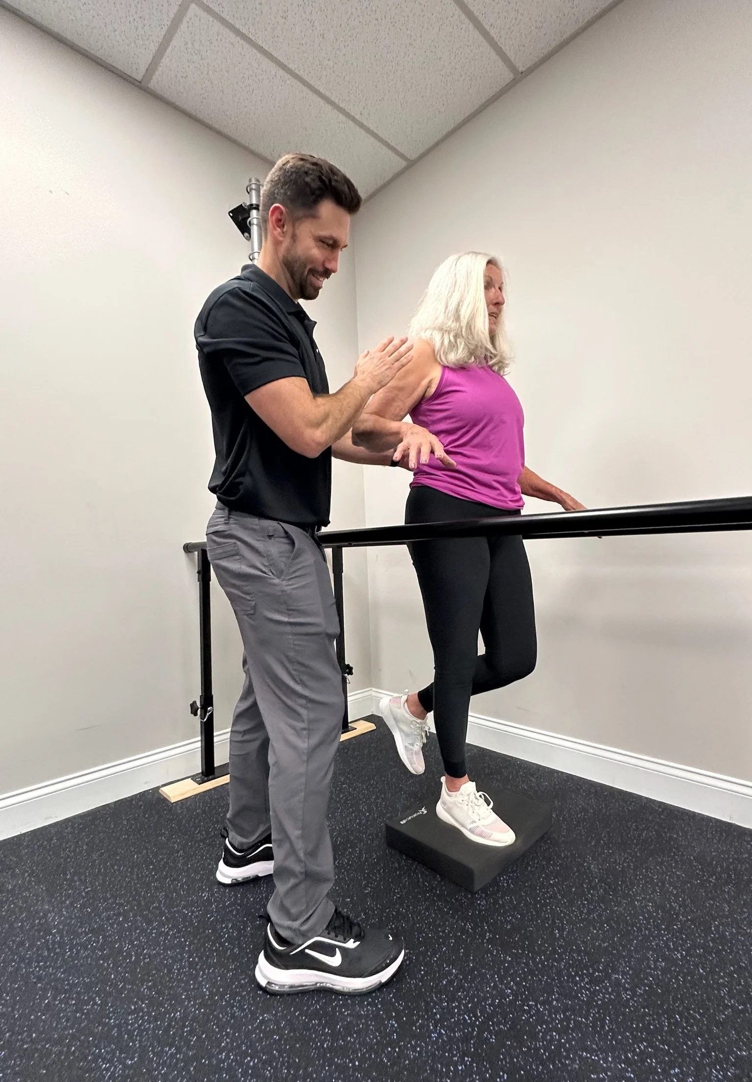 A woman is performing a step exercise on a step platform while being assisted by a physical therapist . The woman is wearing a pink tank top, black leggings, white sneakers, and has blonde hair.