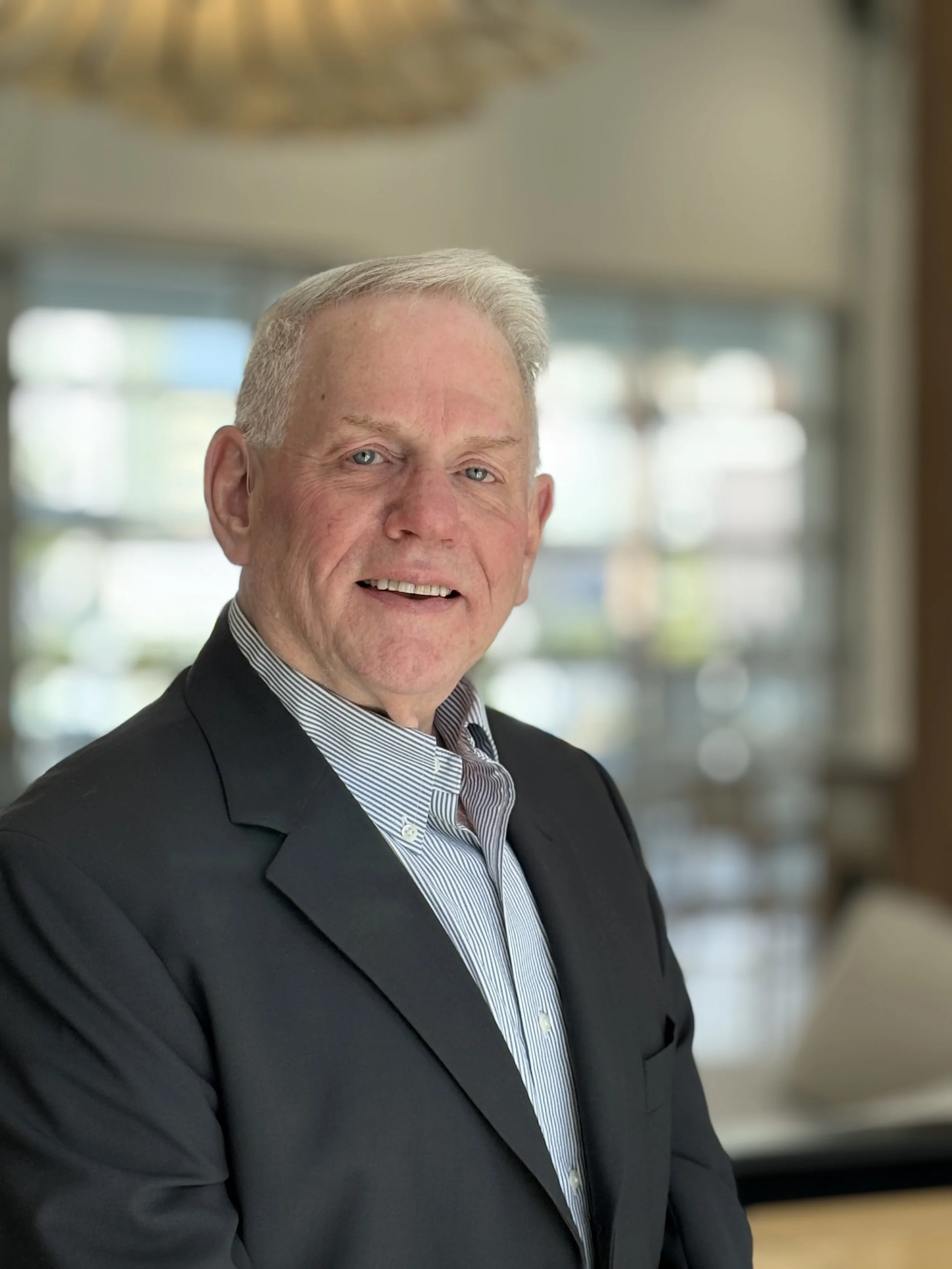 A middle-aged man in a black suit and striped dress shirt smiling in an indoor setting with blurred background windows.