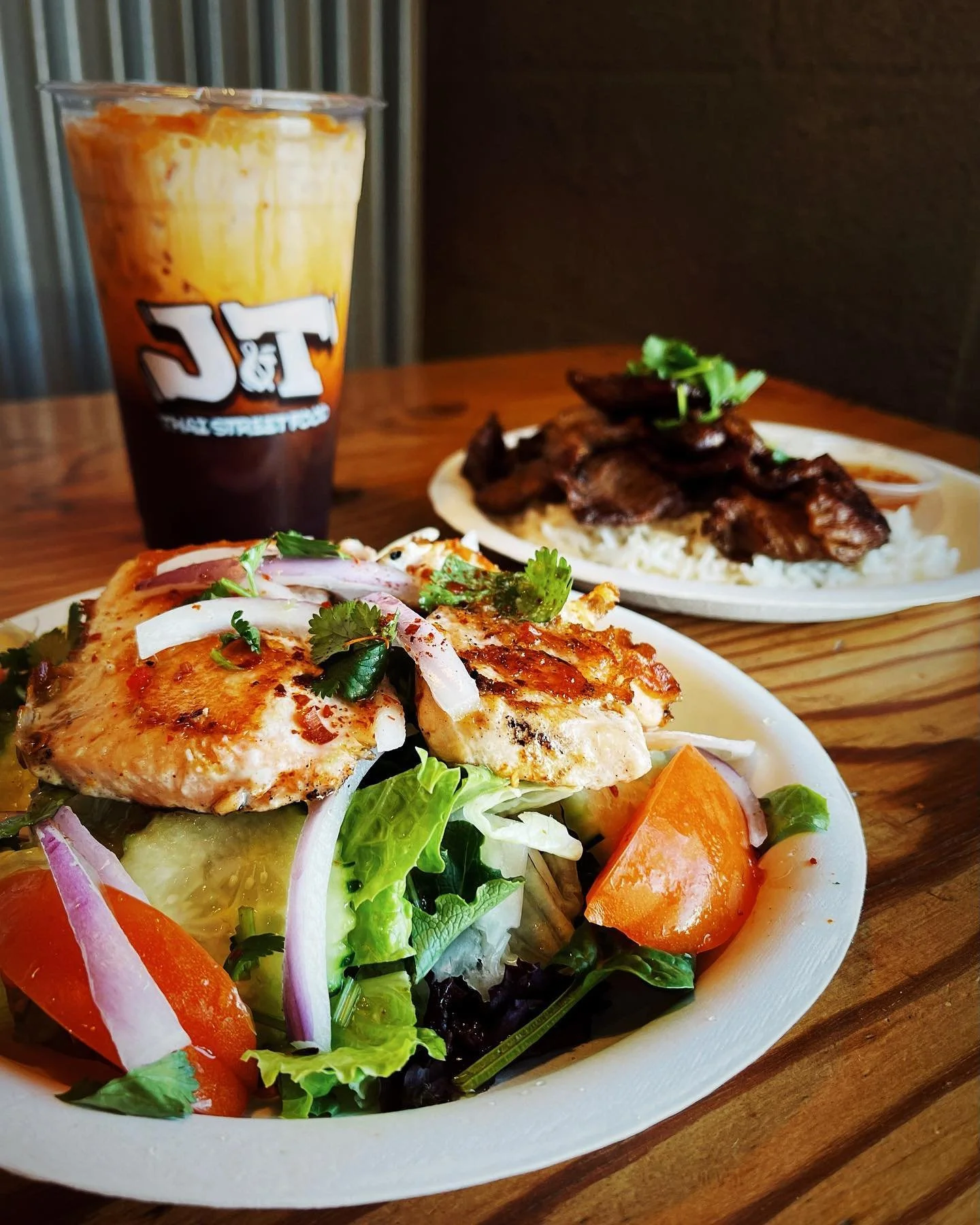 Salad with grilled chicken, vegetables, and herbs, with a plate of beef and rice in the background, and a large iced coffee from J&T.
