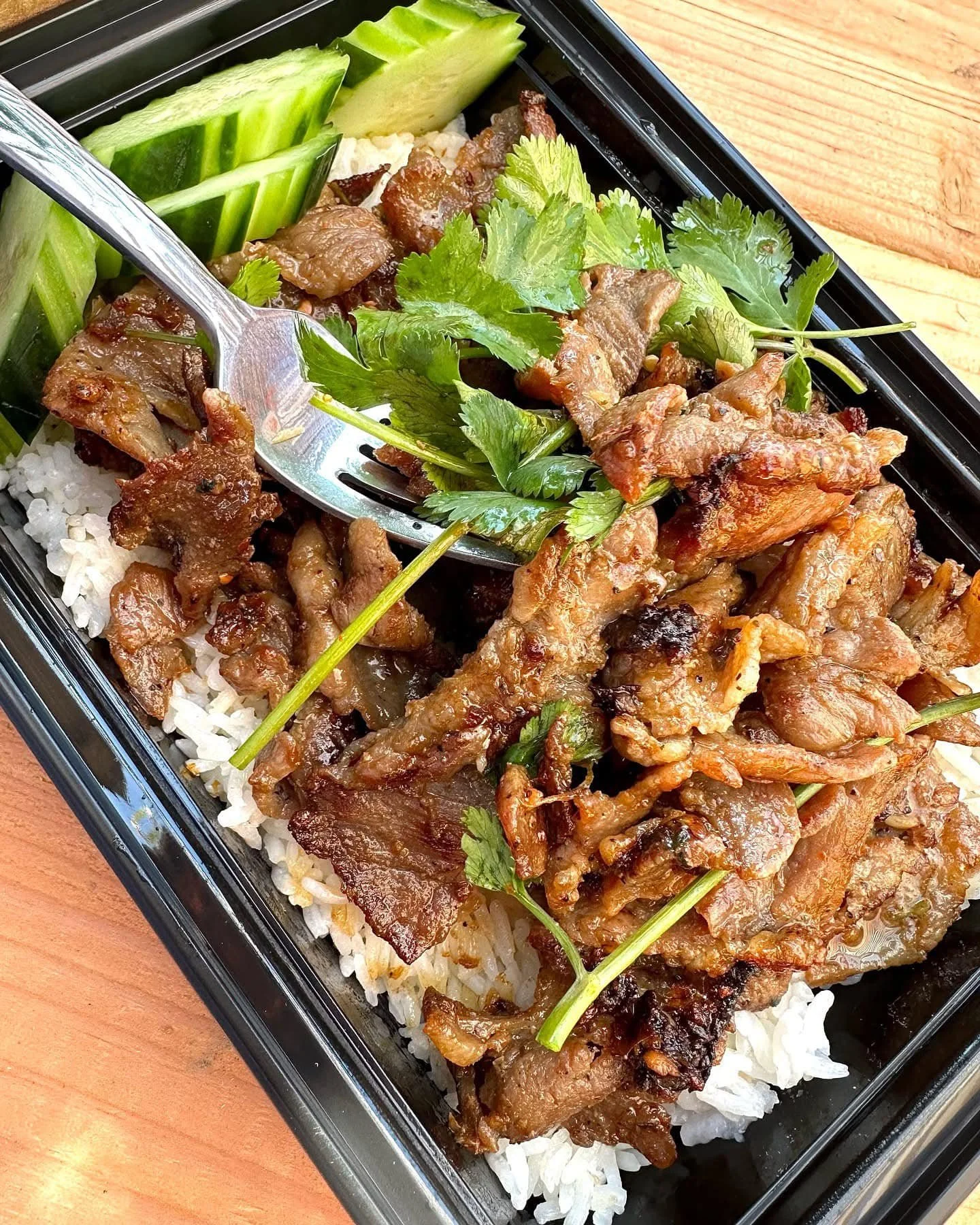 A takeout container filled with grilled beef strips on rice, topped with cilantro, alongside sliced cucumber garnishes.