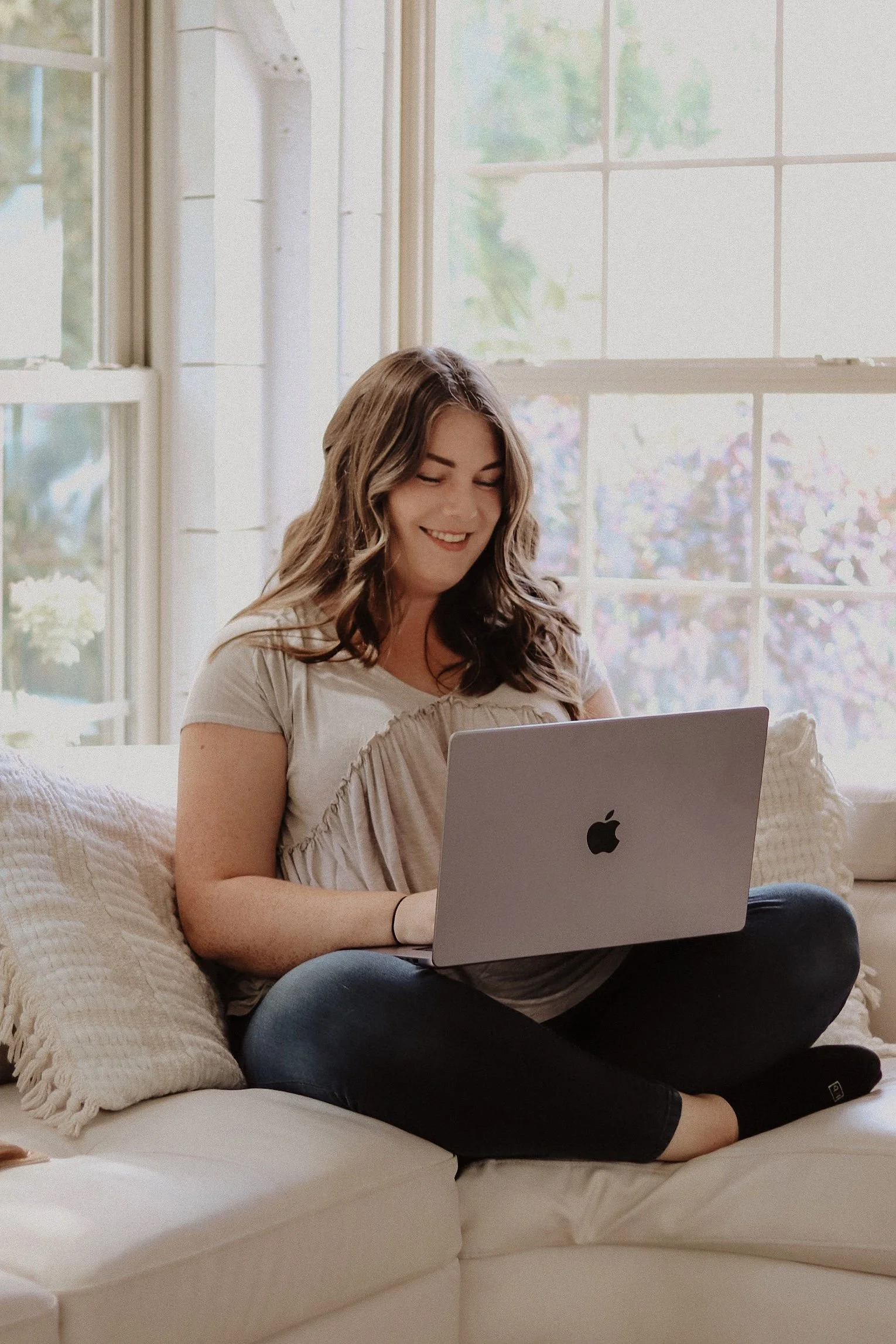 A woman sitting cross-legged on a cream-colored sofa with pillows, using a silver laptop in front of a large window with white frame, in a bright room with natural light.