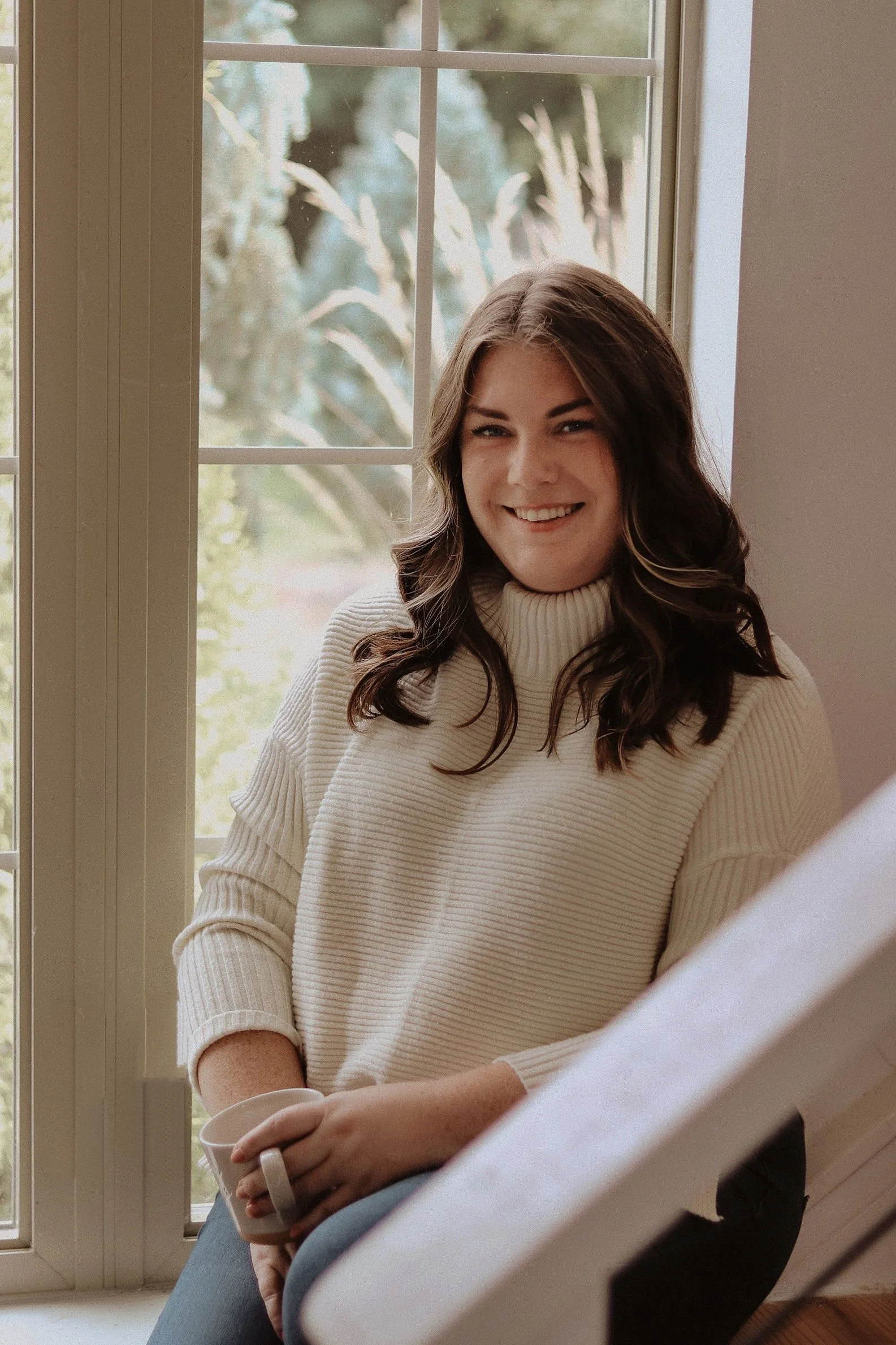 A woman with long, dark, wavy hair sitting by a window, smiling, holding a mug, wearing a cream-colored turtleneck sweater.