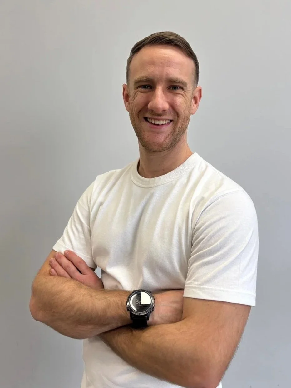 Smiling man with short brown hair, wearing a white t-shirt, standing against a plain gray background, arms crossed, wearing a black watch.