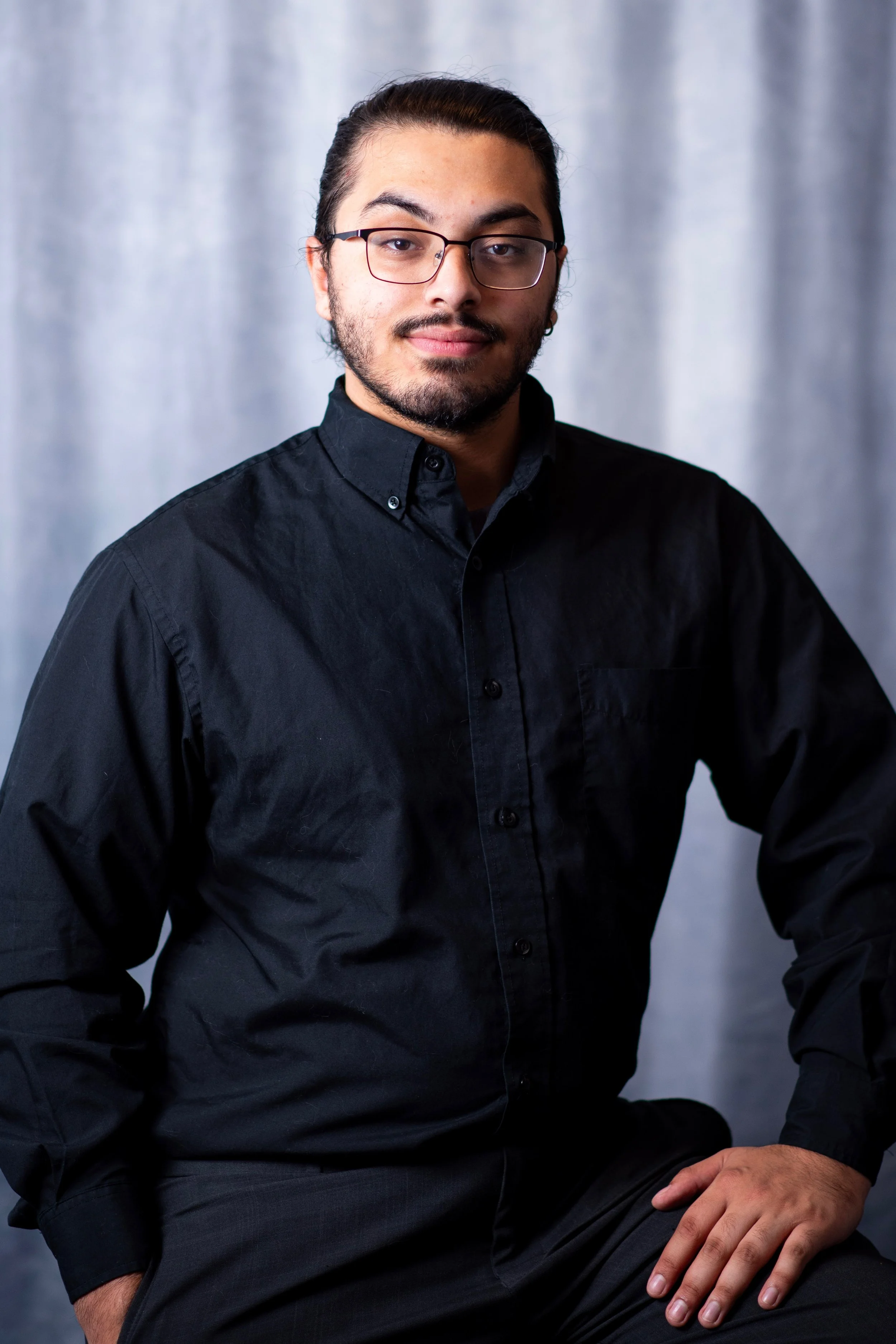 A man with long dark hair, glasses, and a beard wearing a black button-up shirt, seated against a gray background.