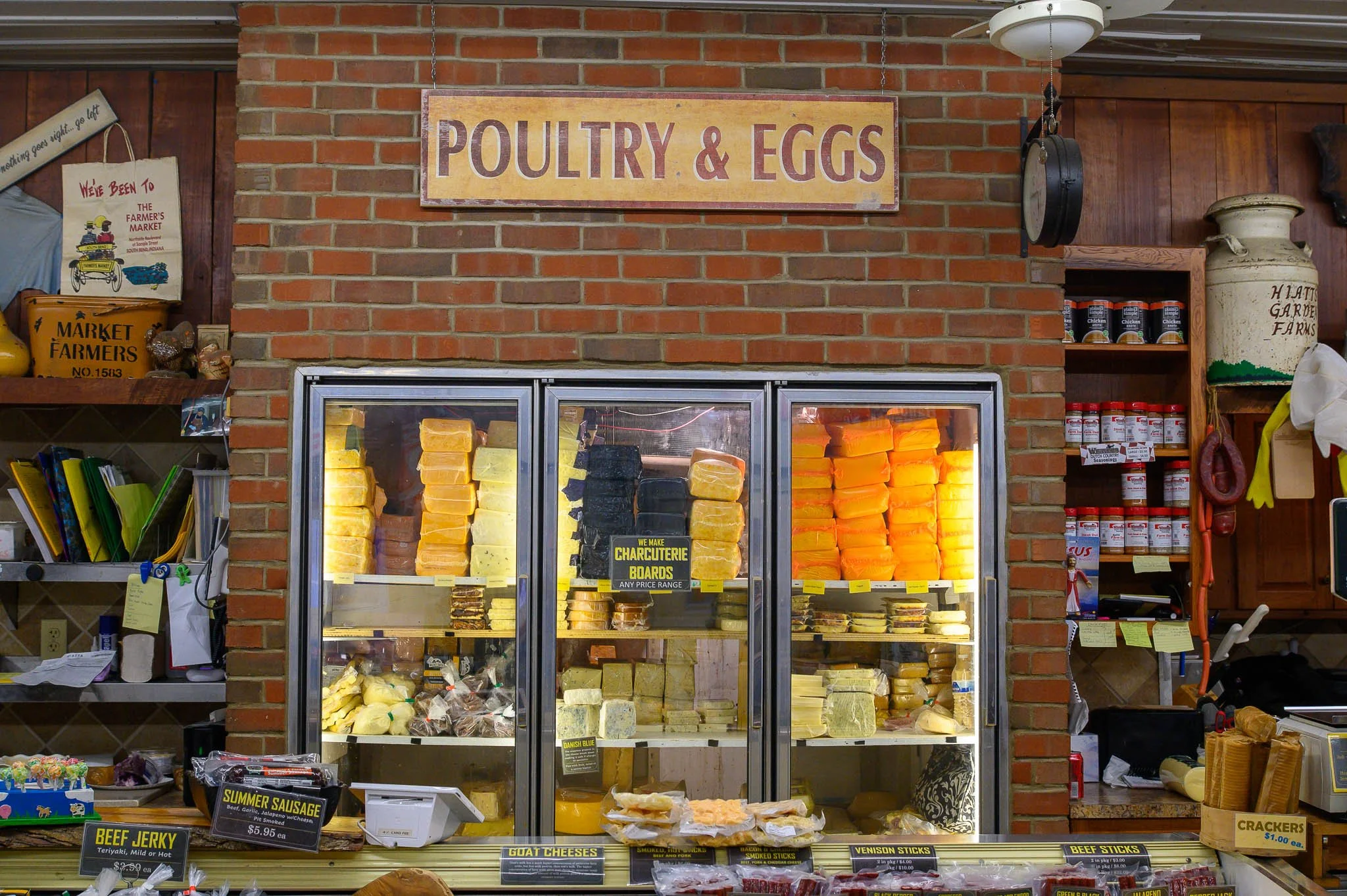Display case filled with various cheeses under a sign reading 'POULTRY & EGGS' inside a brick-walled store. Shelves with jars and packages on either side.