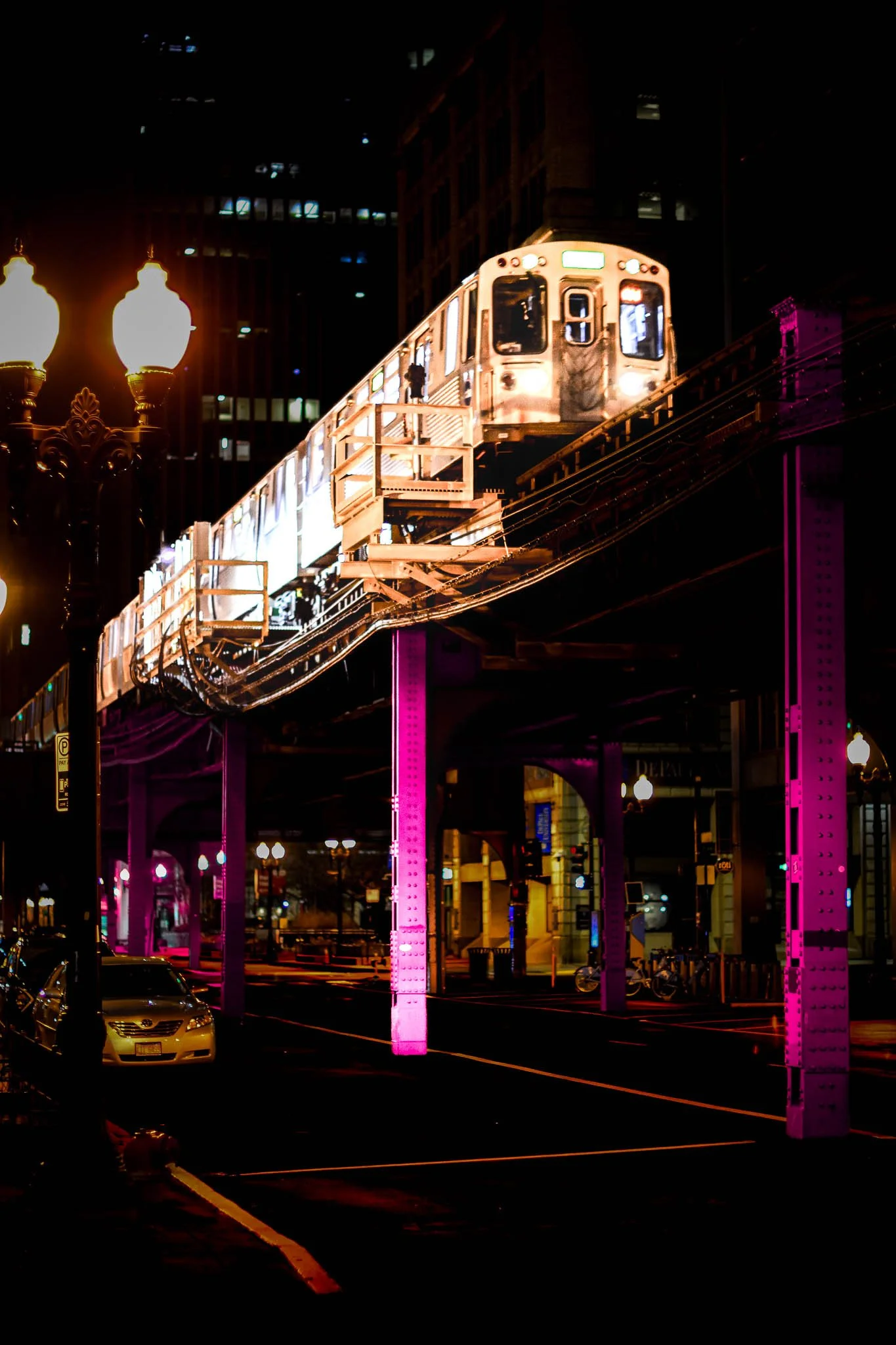 Night scene with an elevated train moving along tracks over a city street illuminated by streetlights and neon purple columns.