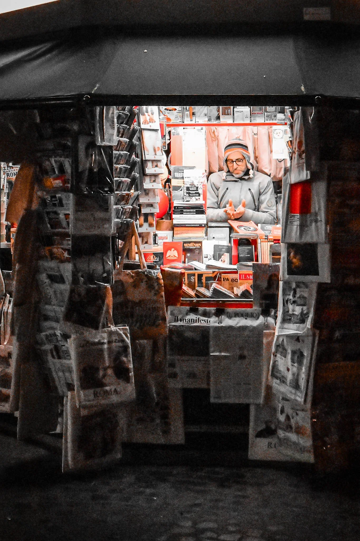 A person standing behind a booth filled with books and magazines, seen through an archway decorated with hanging newspapers.