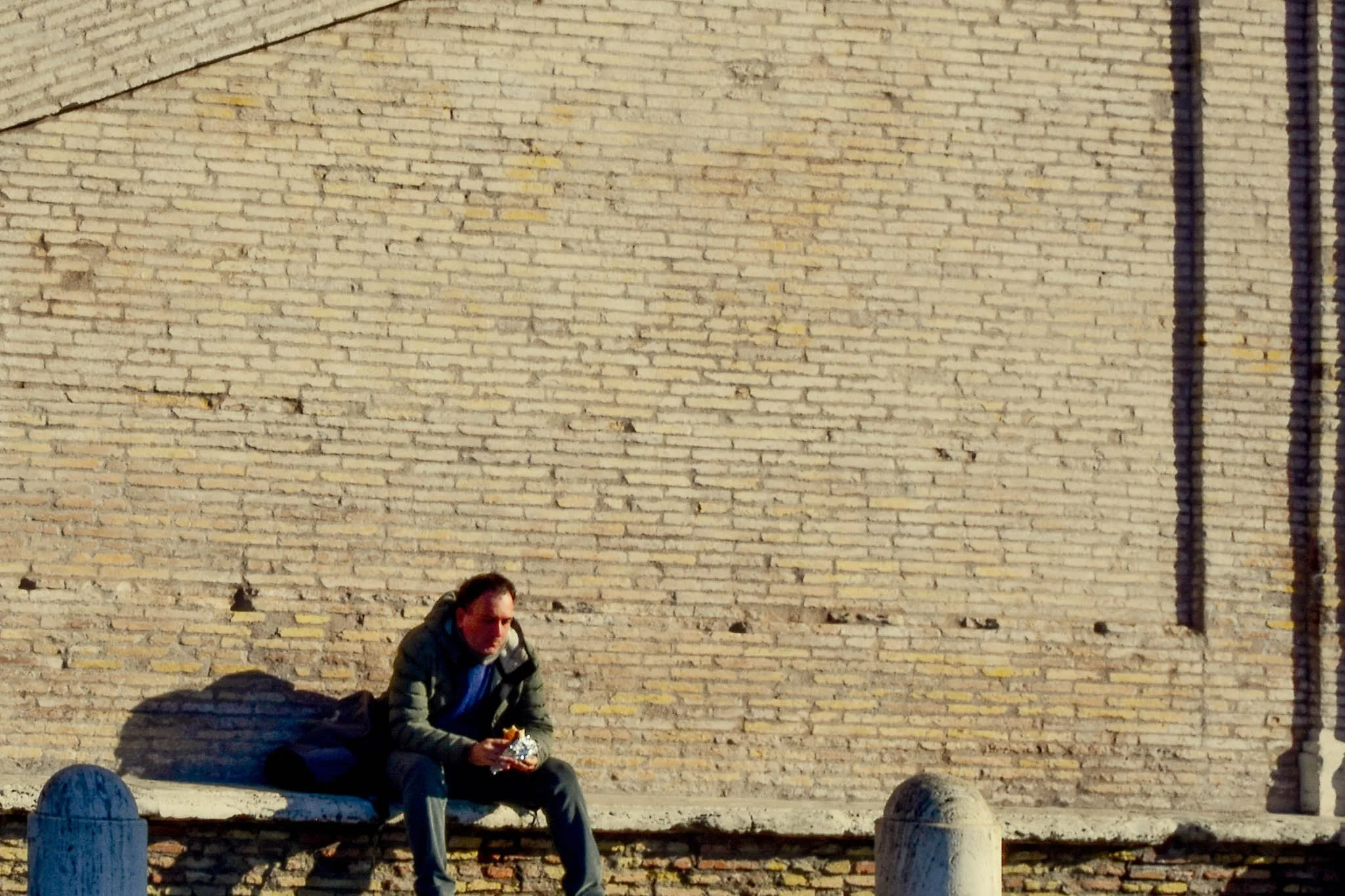 A man sitting on a bench against a large brick wall, eating foil-wrapped food.