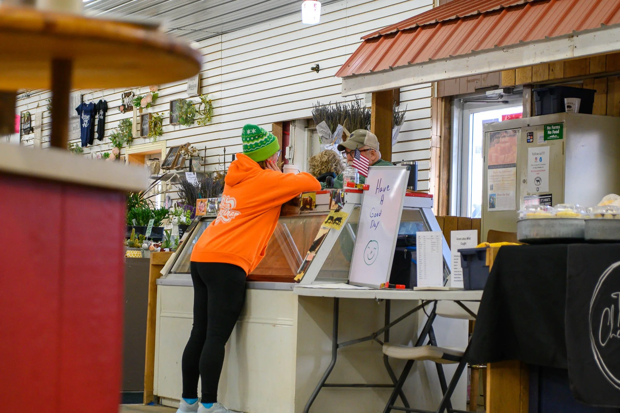 A person in an orange hoodie and green beanie talking to a customer behind a counter at a shop or deli. The customer is an older man wearing sunglasses and a beige cap. The scene is inside a rustic store with white wooden walls and decorative plants.