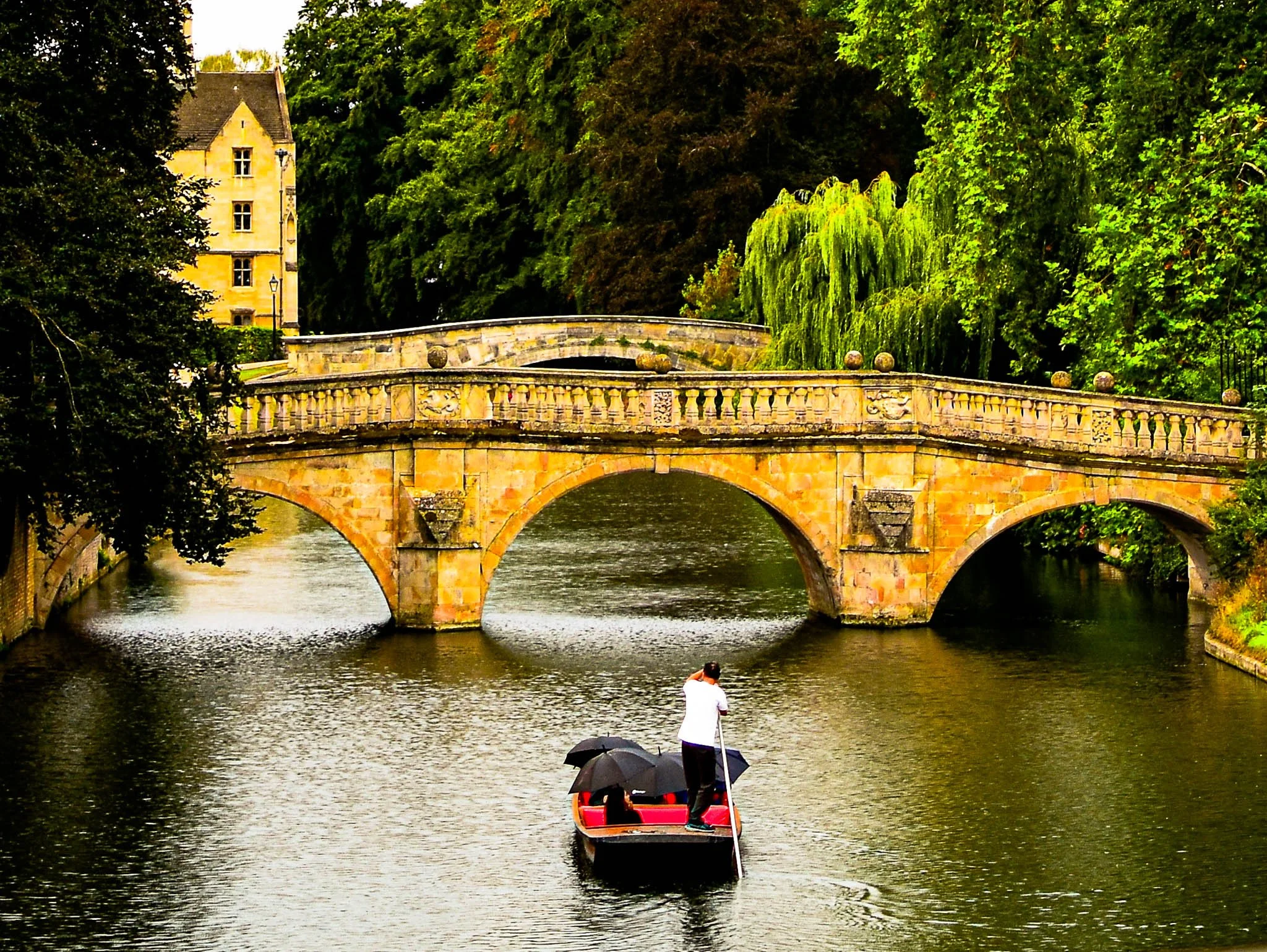 A person standing on a boat with umbrellas, using a pole to navigate a river. In the background, there is a stone bridge and trees along the riverbank.