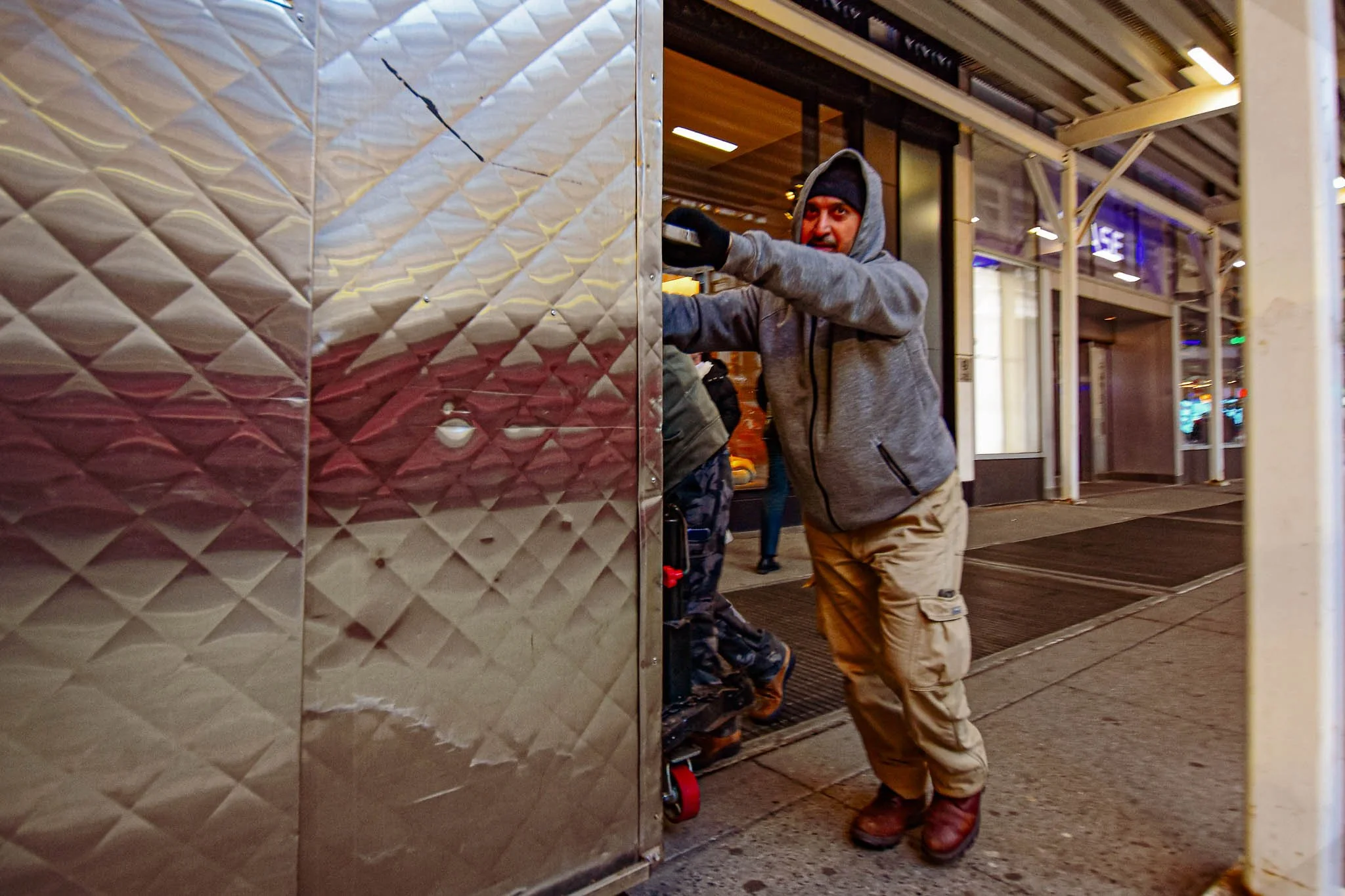 A man in a gray hoodie, black beanie, and tan cargo pants pushing a hand truck through a folding metal door in a city street at night.