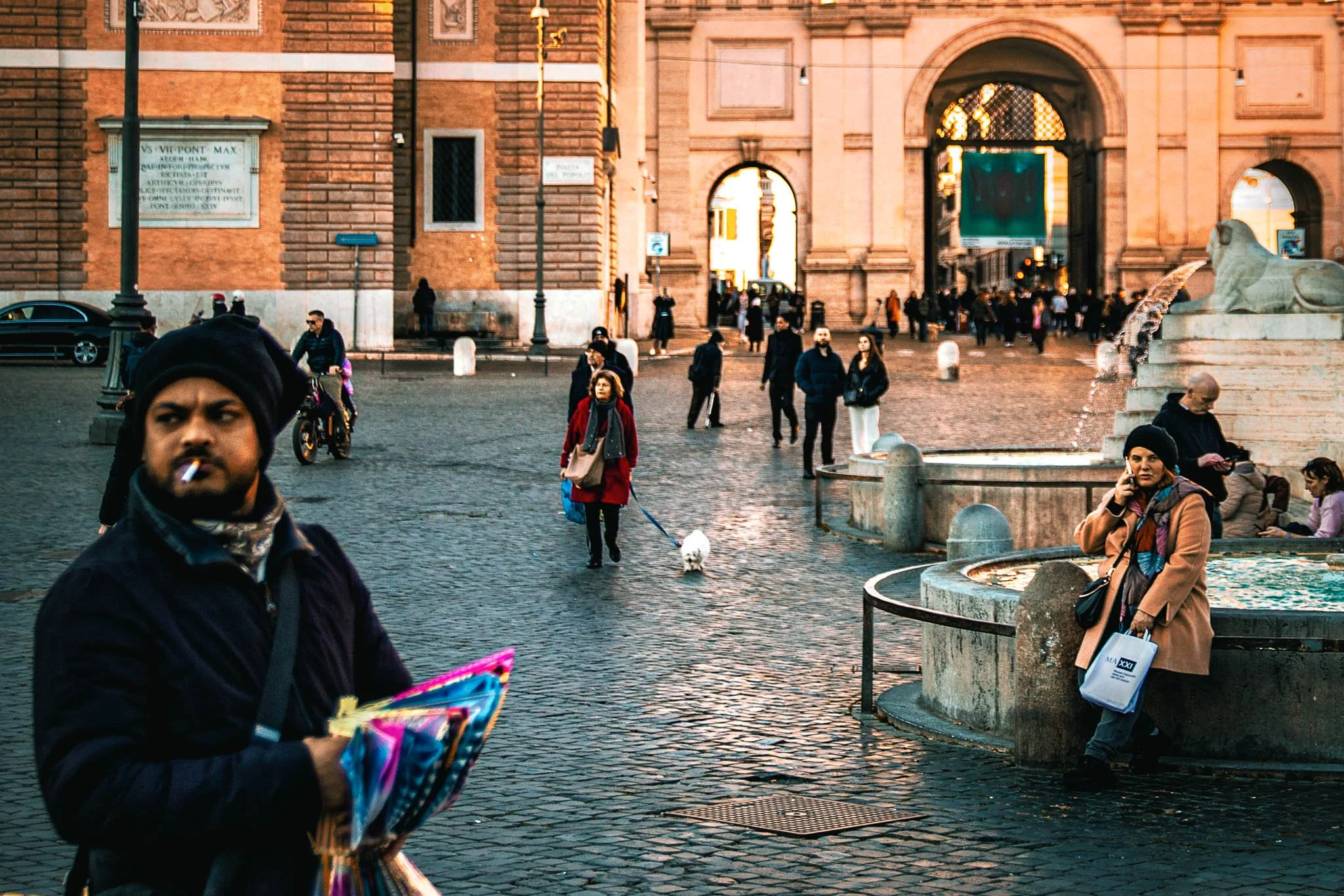 People gather around a fountain at a historic city square with a large arched gateway in the background, in late afternoon or early evening.