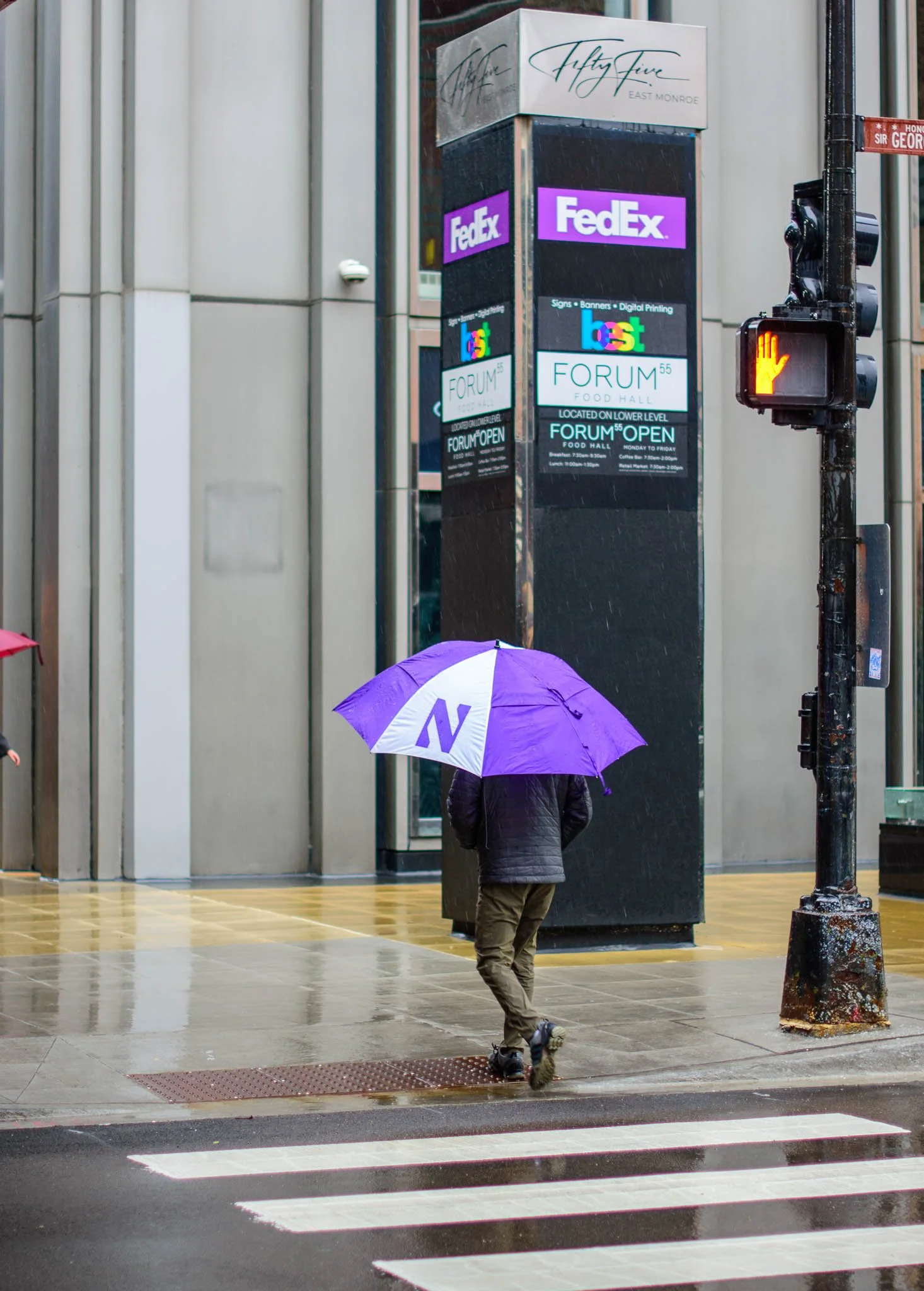 A person walking across a crosswalk in the rain with a purple and white umbrella. They are near a black advertising column with signs for FedEx, Forum, and other businesses, and a hanging traffic signal showing a hand indicating to stop. The scene is