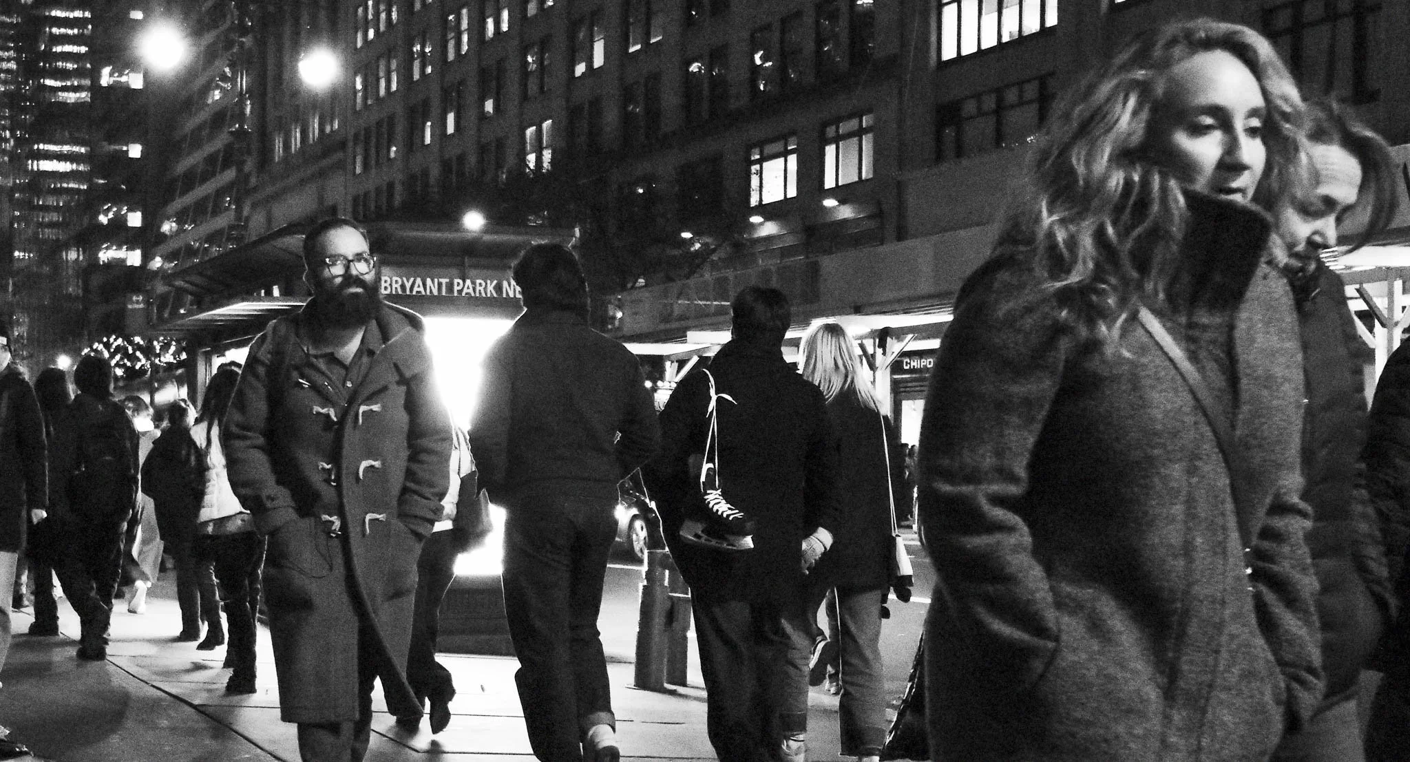 A black-and-white photo of people walking on a city street at night, with a illuminated sign reading 'BRYANT PARK' in the background.