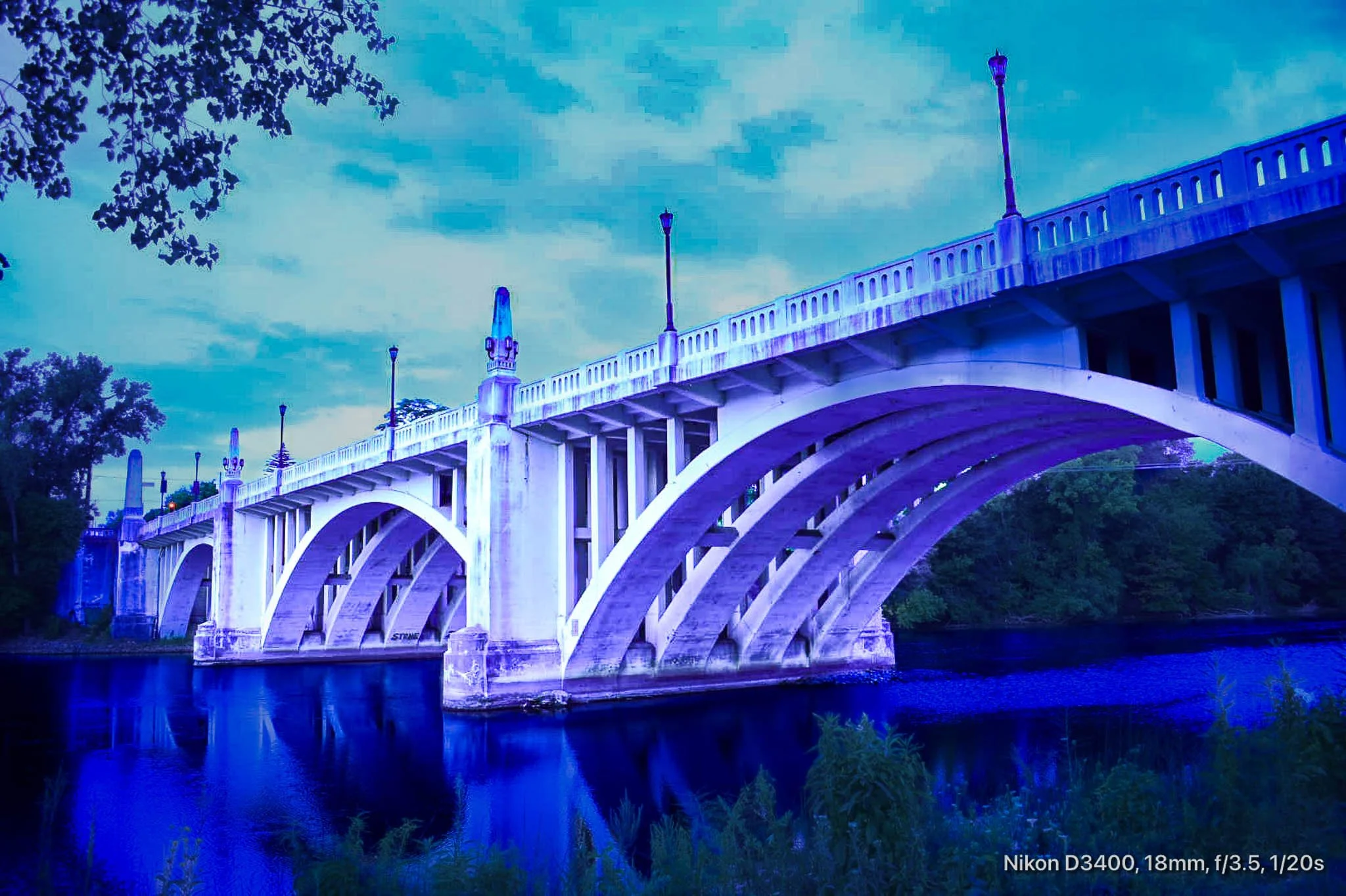 A white arched bridge over a river, with street lamps along the top, under a cloudy sky in a natural setting.