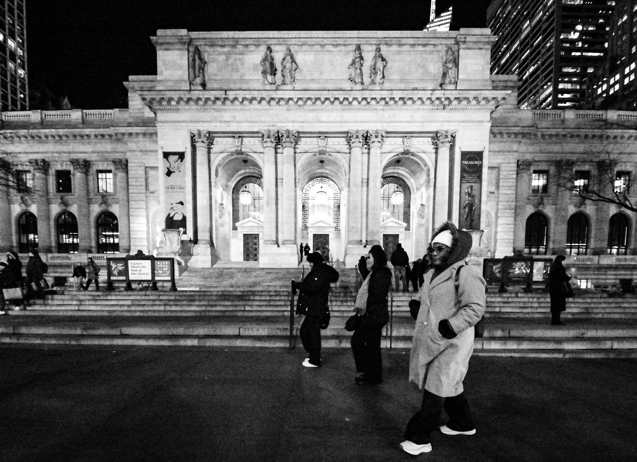 Nighttime scene of a group of people walking down stairs in front of a grand neoclassical building with columns and sculptures.