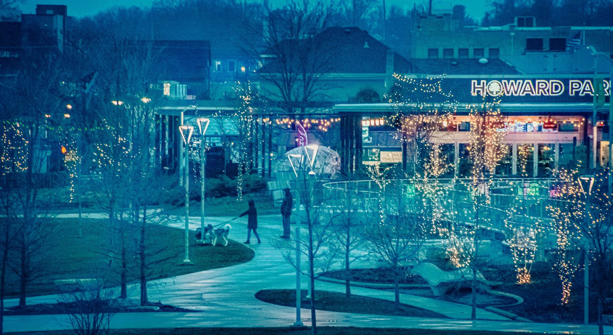 Nighttime scene of a park with leafless trees decorated with string lights, a winding walkway, people walking a dog, and a building in the background with the sign 'Howard PA' and festive lights.