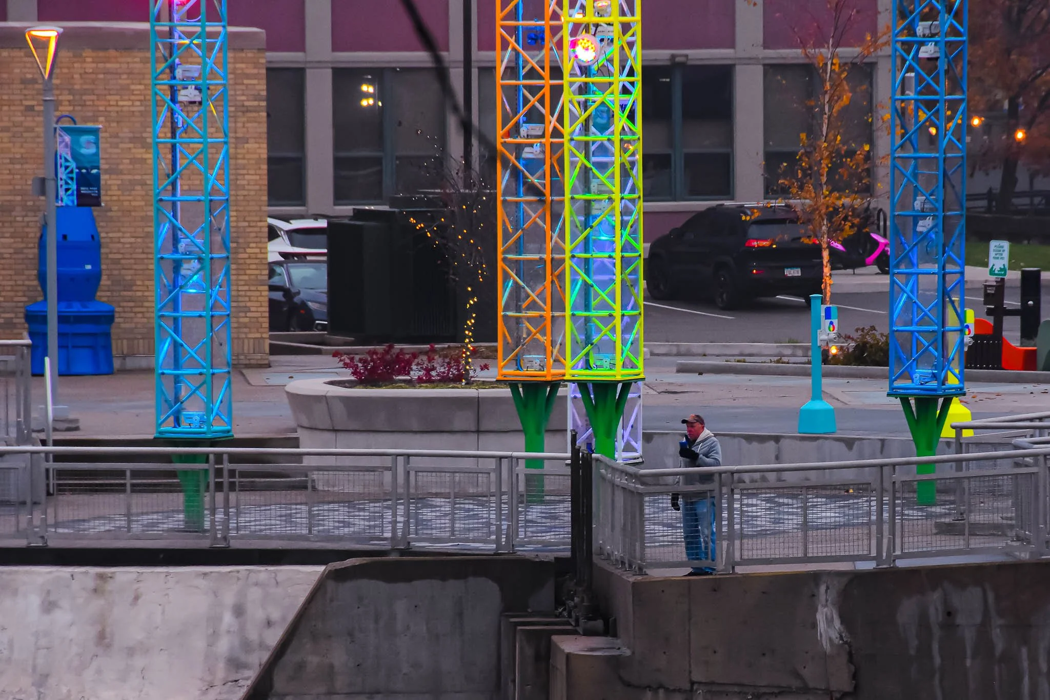 Person standing on a bridge in an urban area, looking at their phone, with colorful metallic structures and a little snow on the ground.
