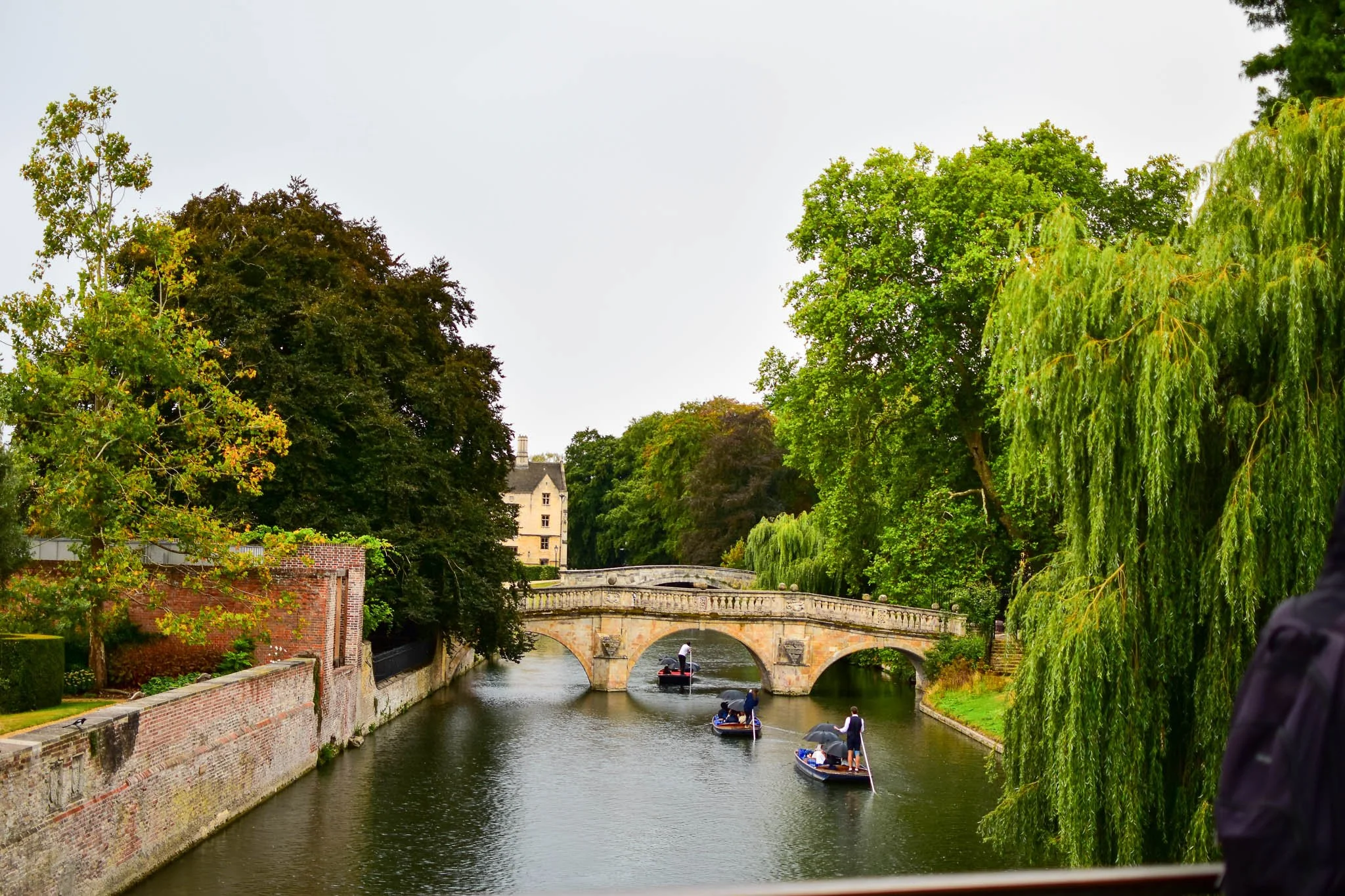 View of a river with a stone bridge, surrounded by lush green trees, and boats with people on the water.