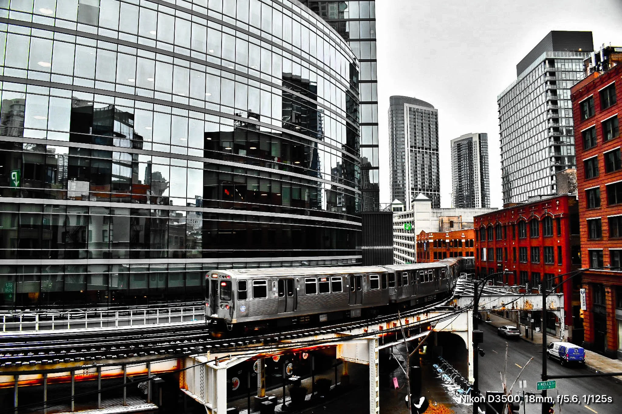 A silver train travels on elevated tracks through a city with modern glass buildings and red brick buildings.