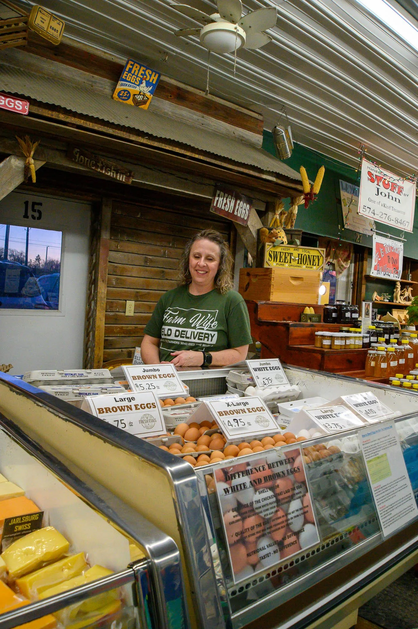 Woman selling eggs at a farm stand, with signs for fresh eggs, honey, and other farm products, inside a rustic wooden building.