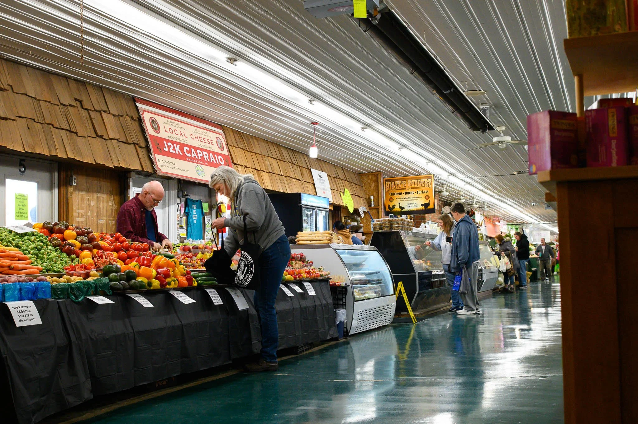 Indoor market with fresh produce, including tomatoes, carrots, and peppers, and customers shopping at stalls with signs for local cheese and poultry.