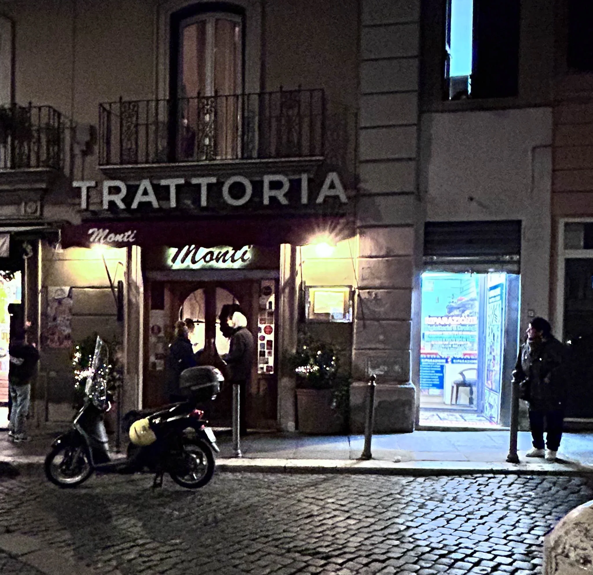 Nighttime street scene with a trattoria restaurant, people waiting outside, a scooter parked on cobblestone street, and a small illuminated shop entrance.
