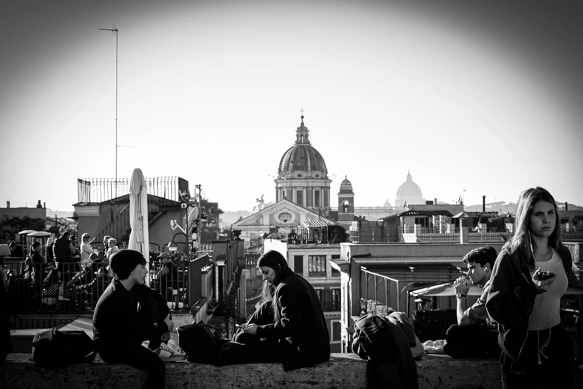Group of young people sitting and standing on a rooftop terrace with city buildings and domes in the background, black and white photo.